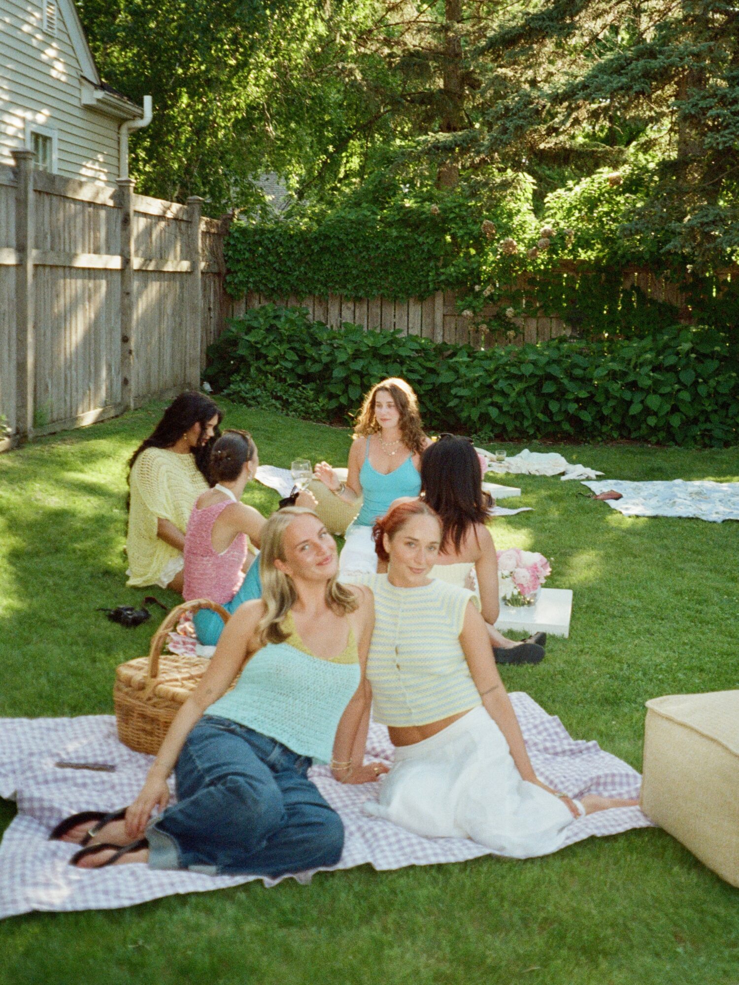 Group of women and children sitting on a blanket in a backyard with trees and a wooden fence.
