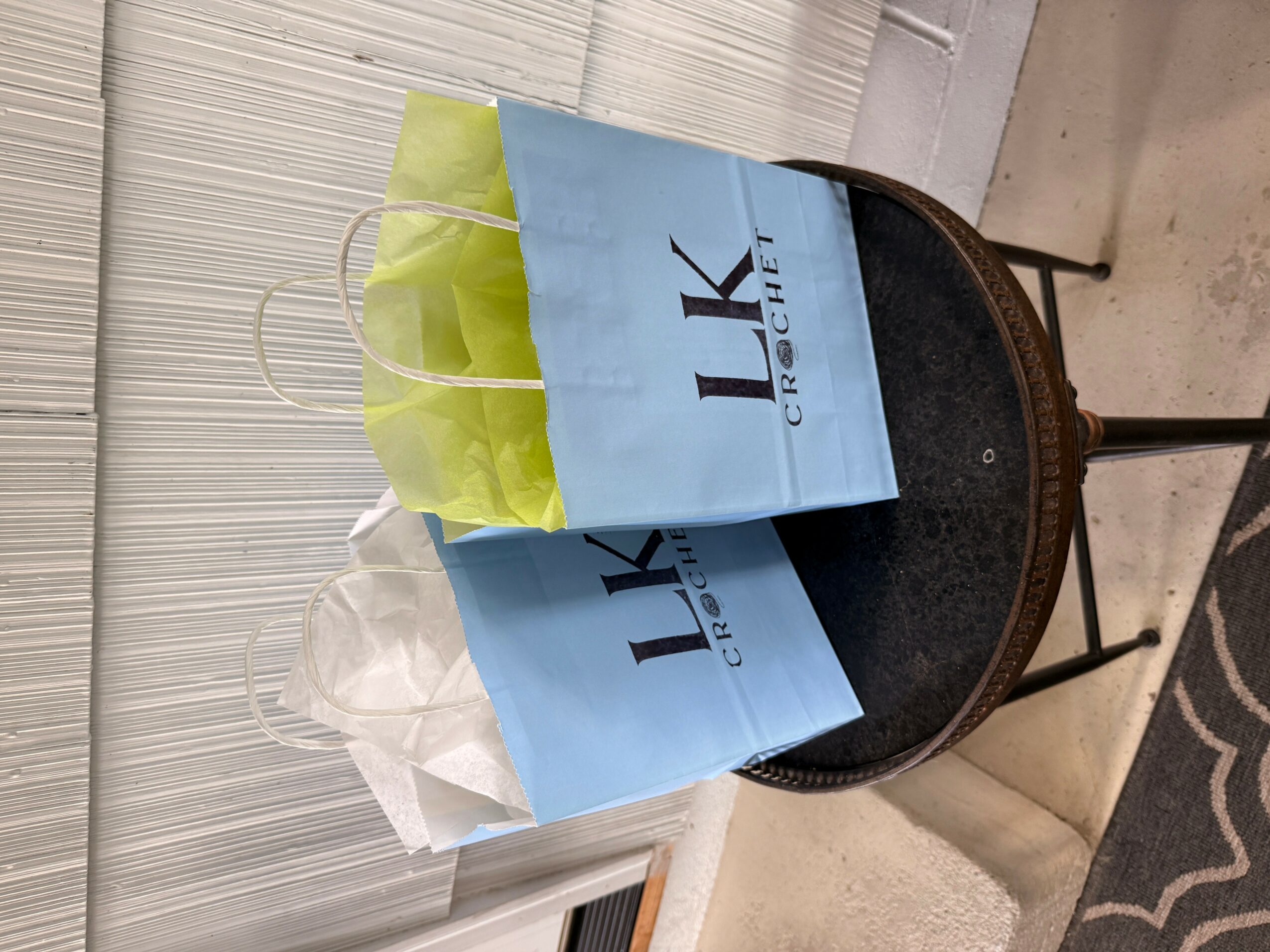 Two shopping bags, one green and one white, placed on a black chair with a blue bag behind them, against a wall.