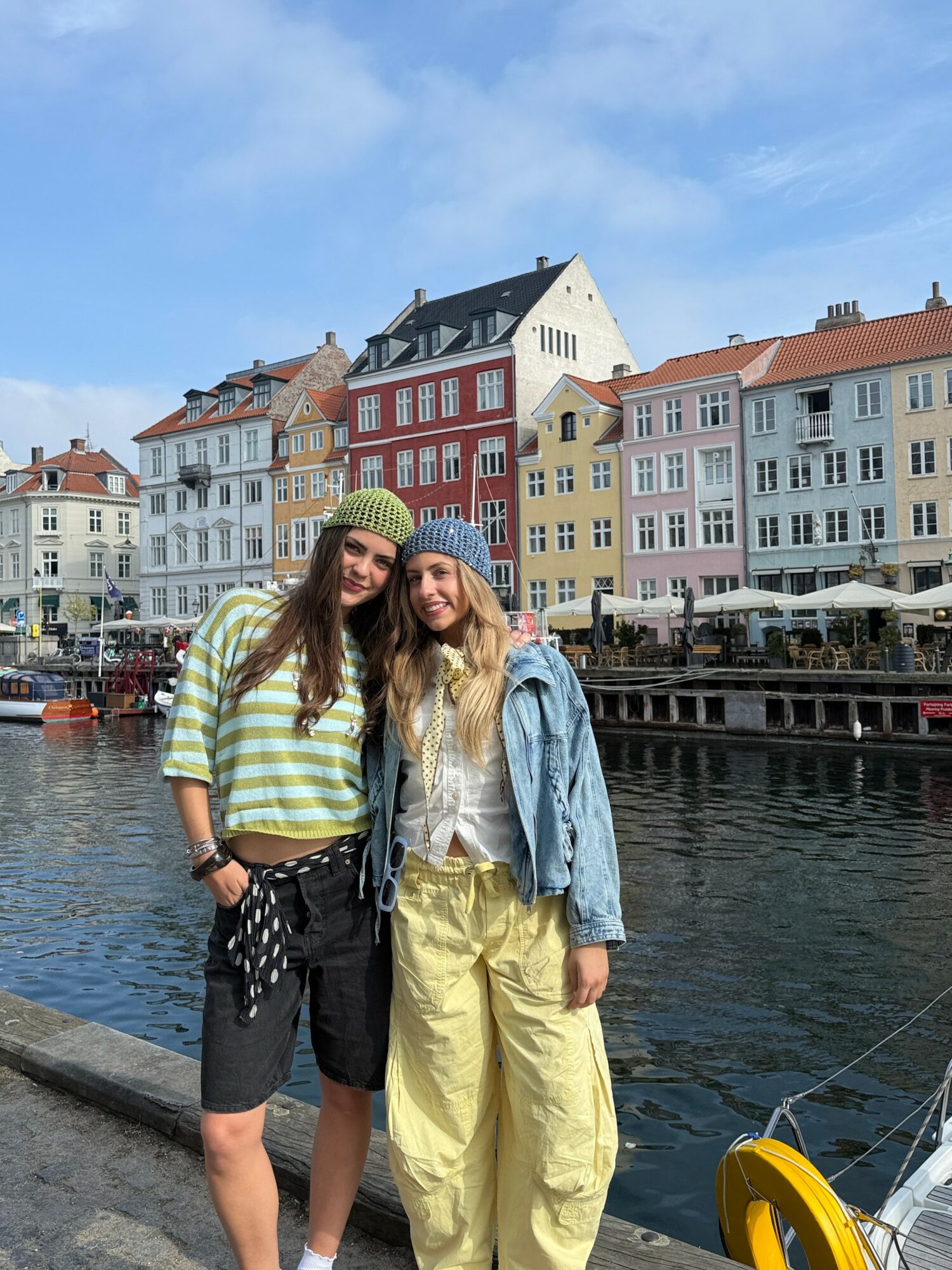 Two women stand by a canal with colorful buildings in the background, smiling and wearing casual clothing and knit hats.