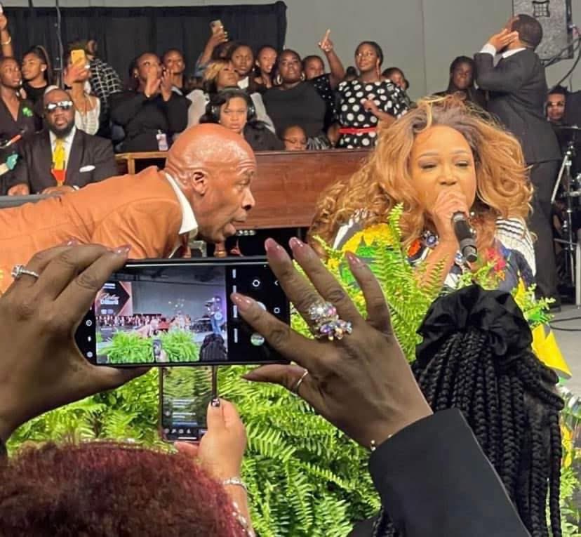 A woman with long, curly hair sings into a microphone, surrounded by people, with a man leaning in close, and audience in background.