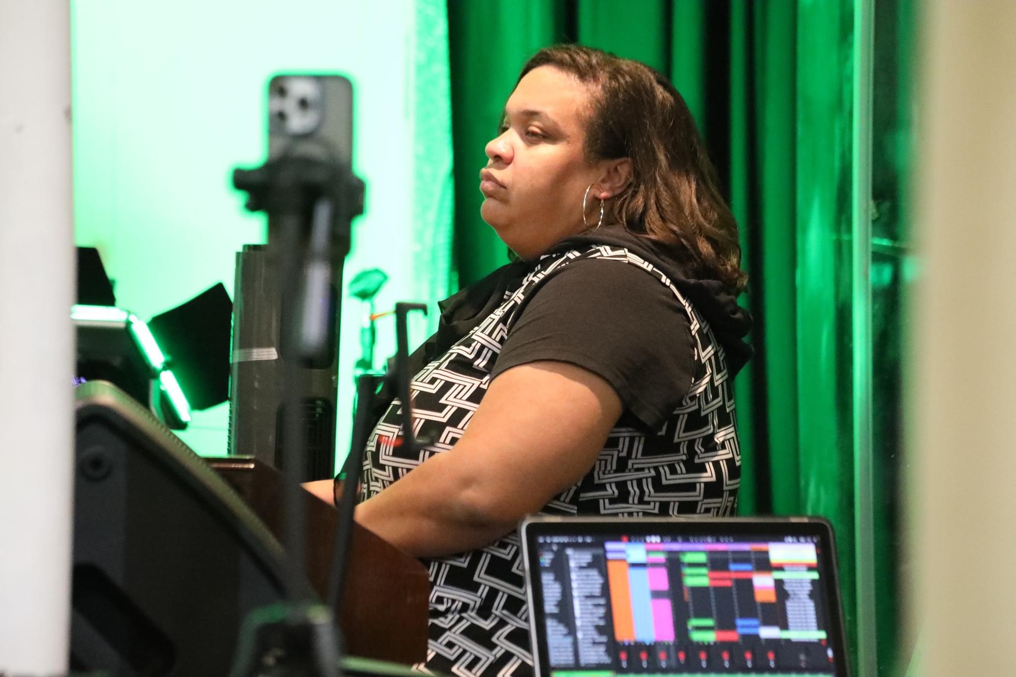 Woman with shoulder-length hair sitting in front of a computer with a colorful screen, green curtains in the background.