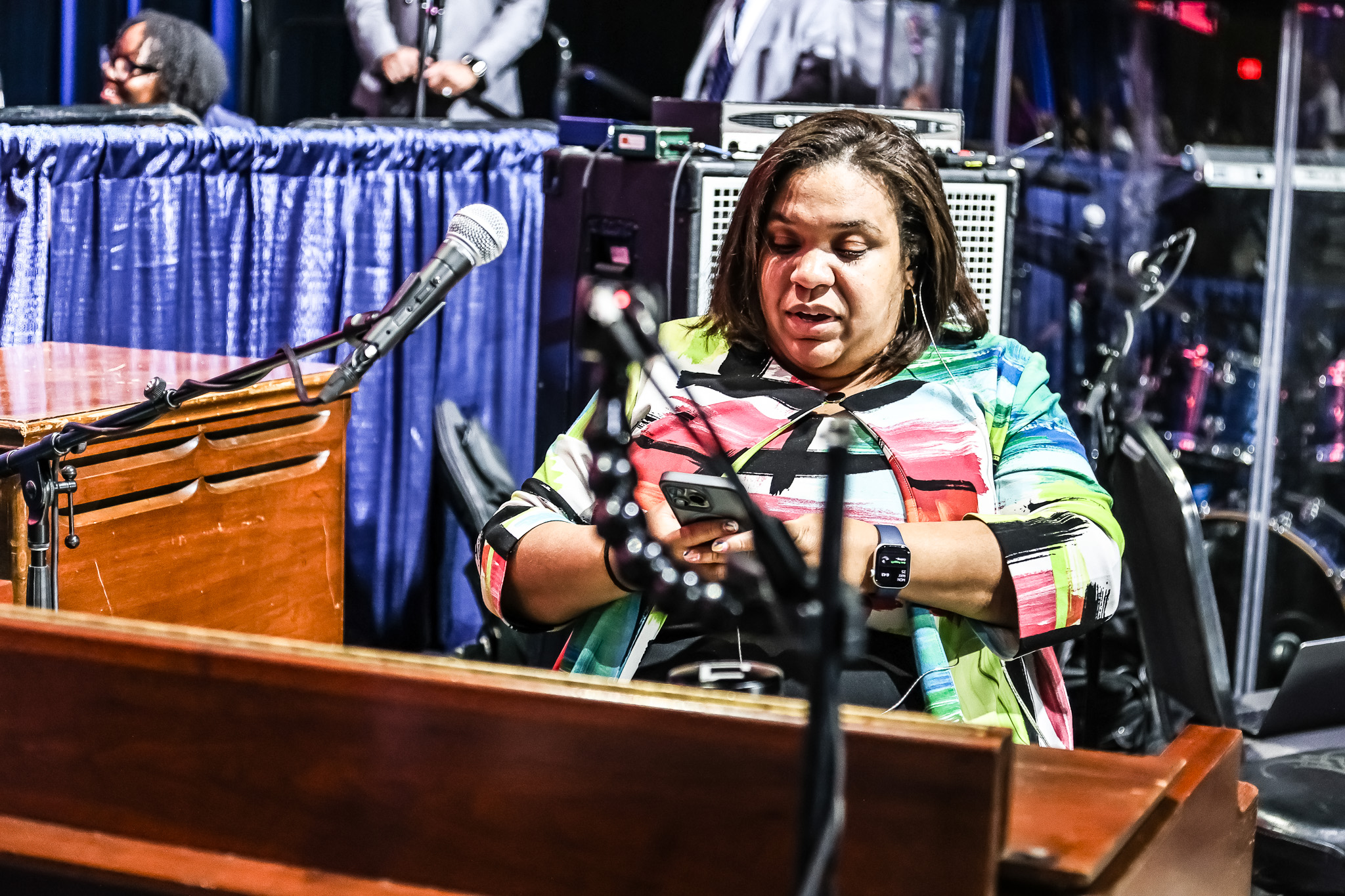 Woman sitting at a table with a microphone, looking at her phone, surrounded by audio equipment and a blue curtain.
