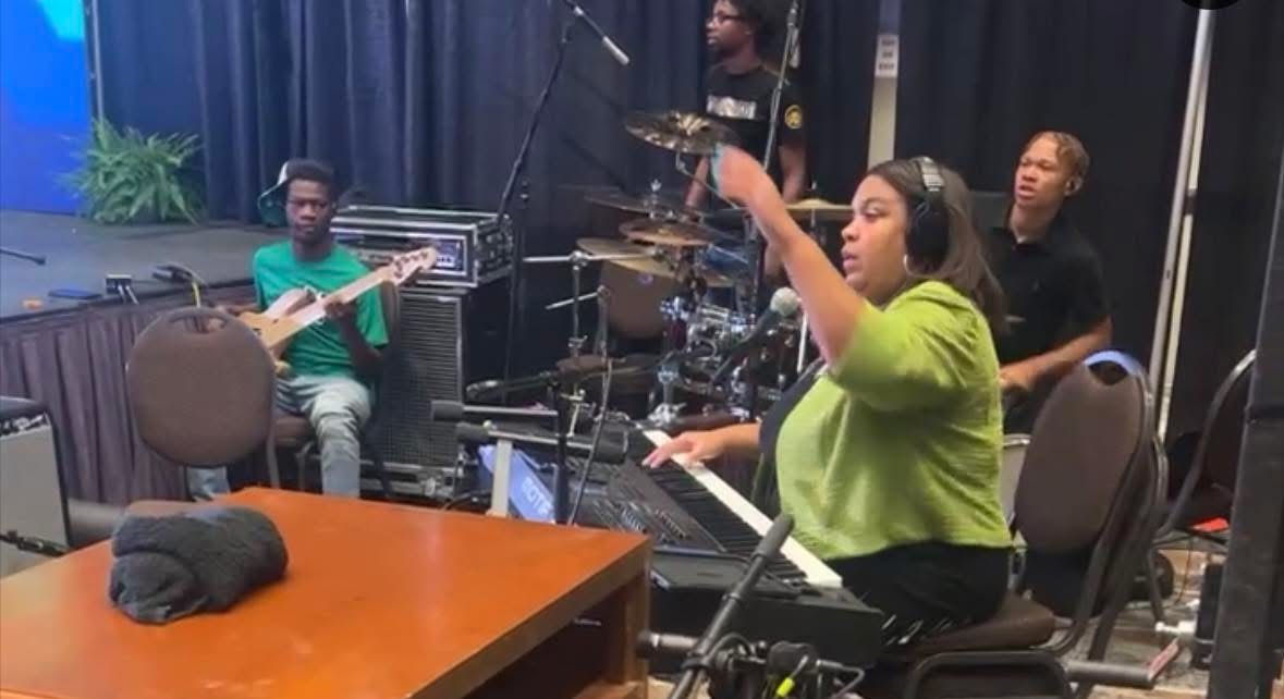 Group of four people in a music rehearsal room, one playing keyboard, others with instruments, woman pointing, table in foreground.