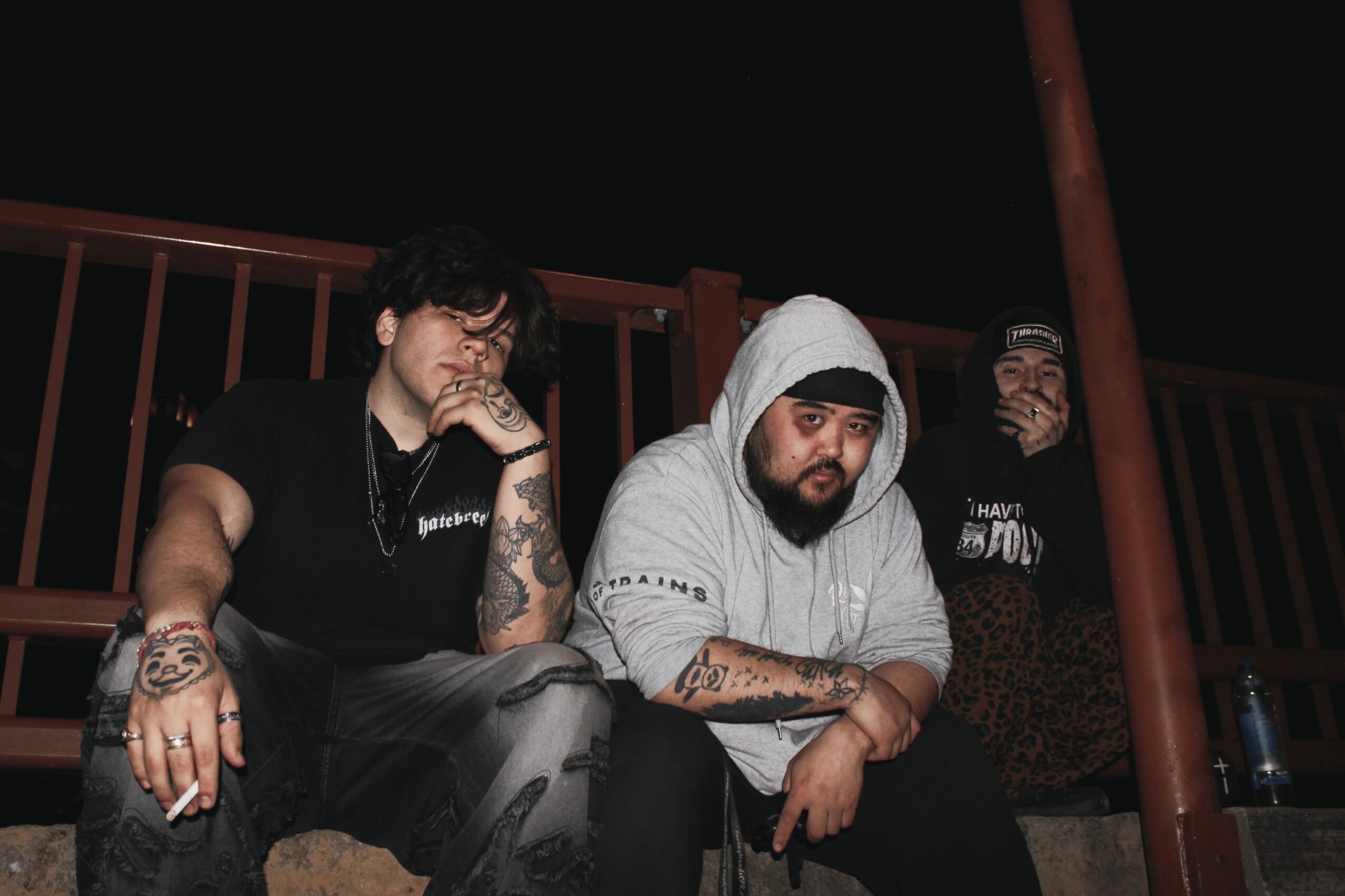 Three men sitting outdoors at night, one wearing a hoodie, two with tattoos, behind a railing, with bottles nearby.