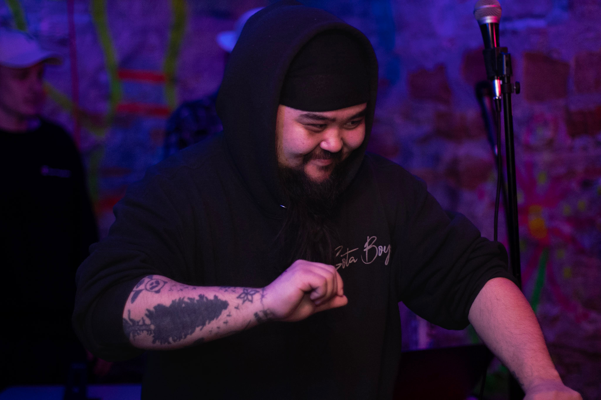 Person with a beard and tattooed arm smiling, wearing a black hoodie, in a colorful, dimly lit setting.