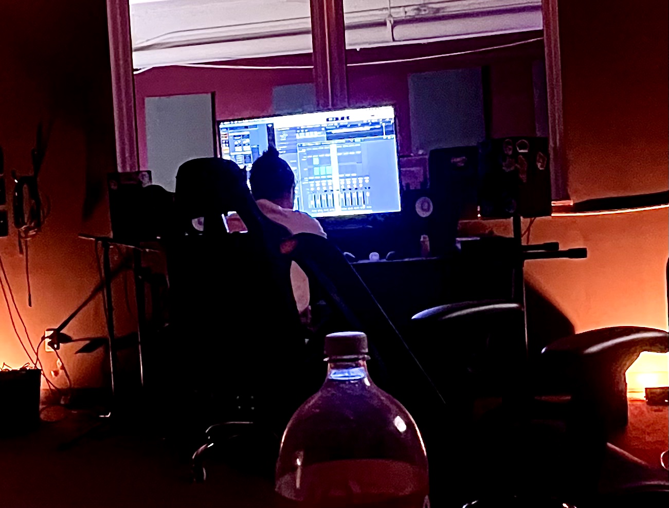 Person working at a computer in a dimly lit room with orange walls, music studio equipment, and a water bottle in foreground.