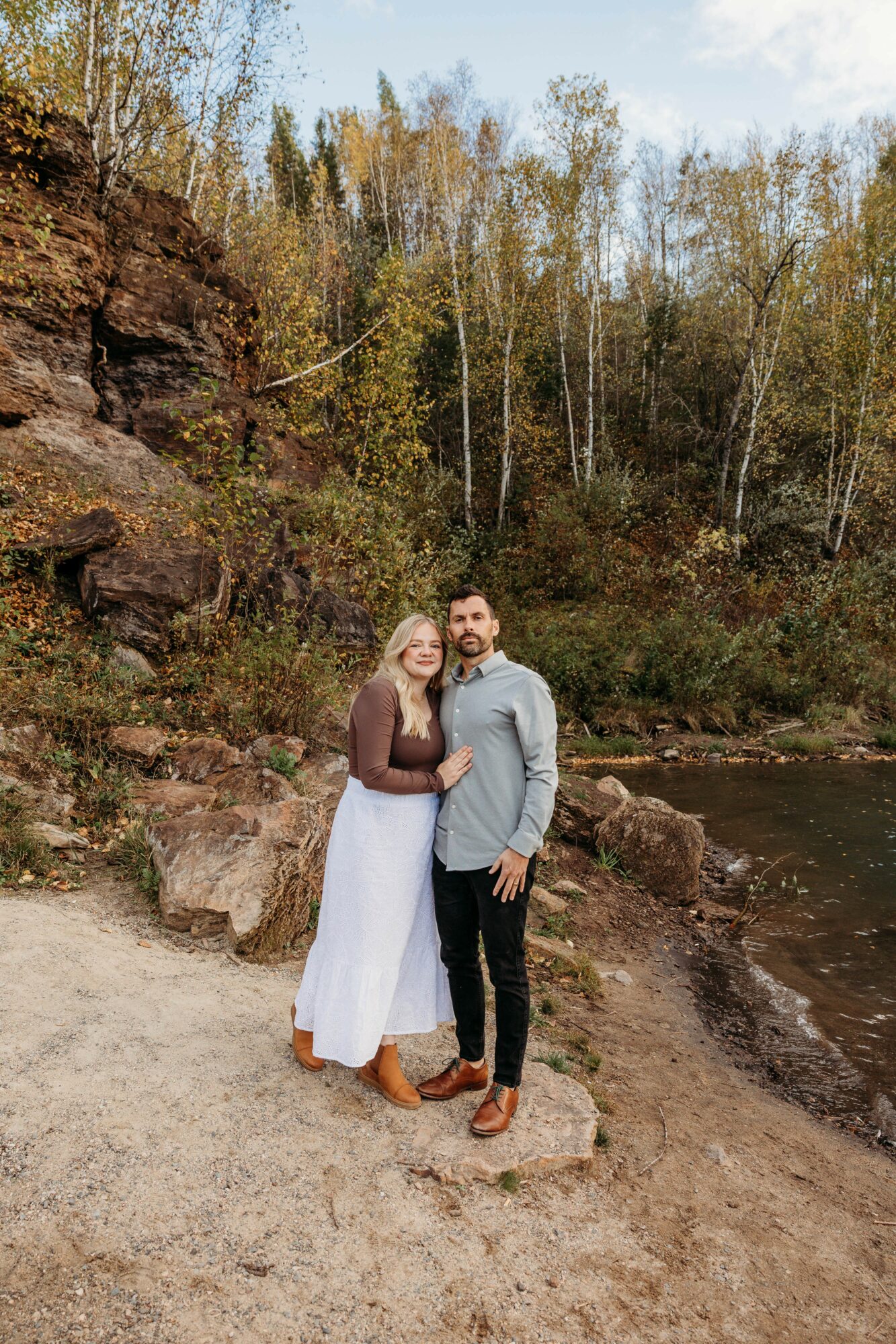 A couple stands on a rocky riverbank with trees and a river in the background.