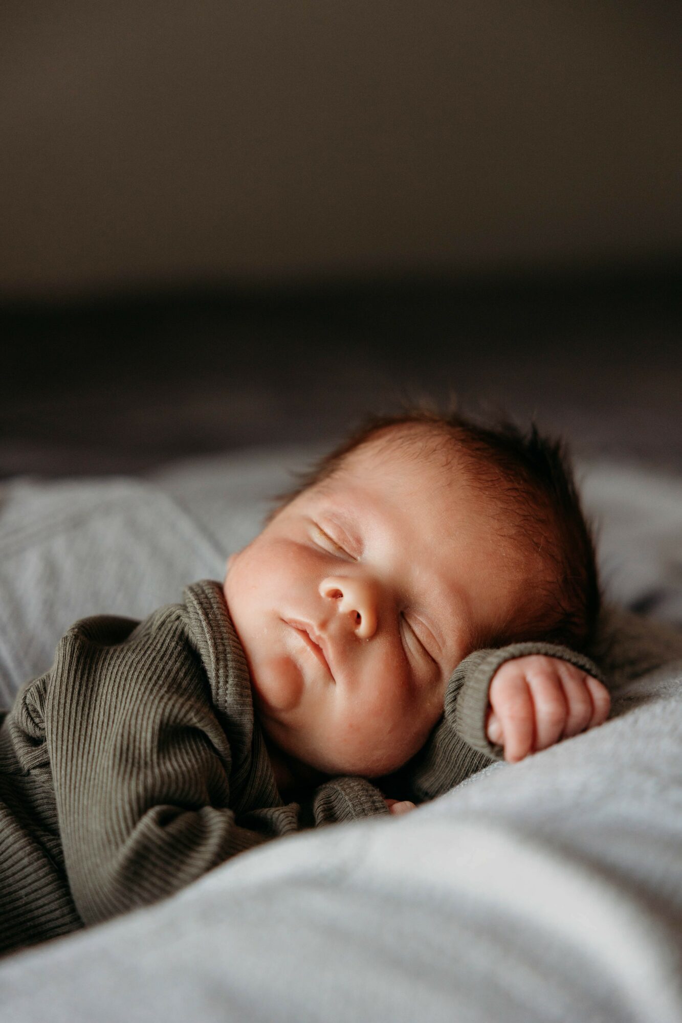 Baby sleeping peacefully on a soft surface, wearing a dark green outfit, with eyes closed and hand near face.