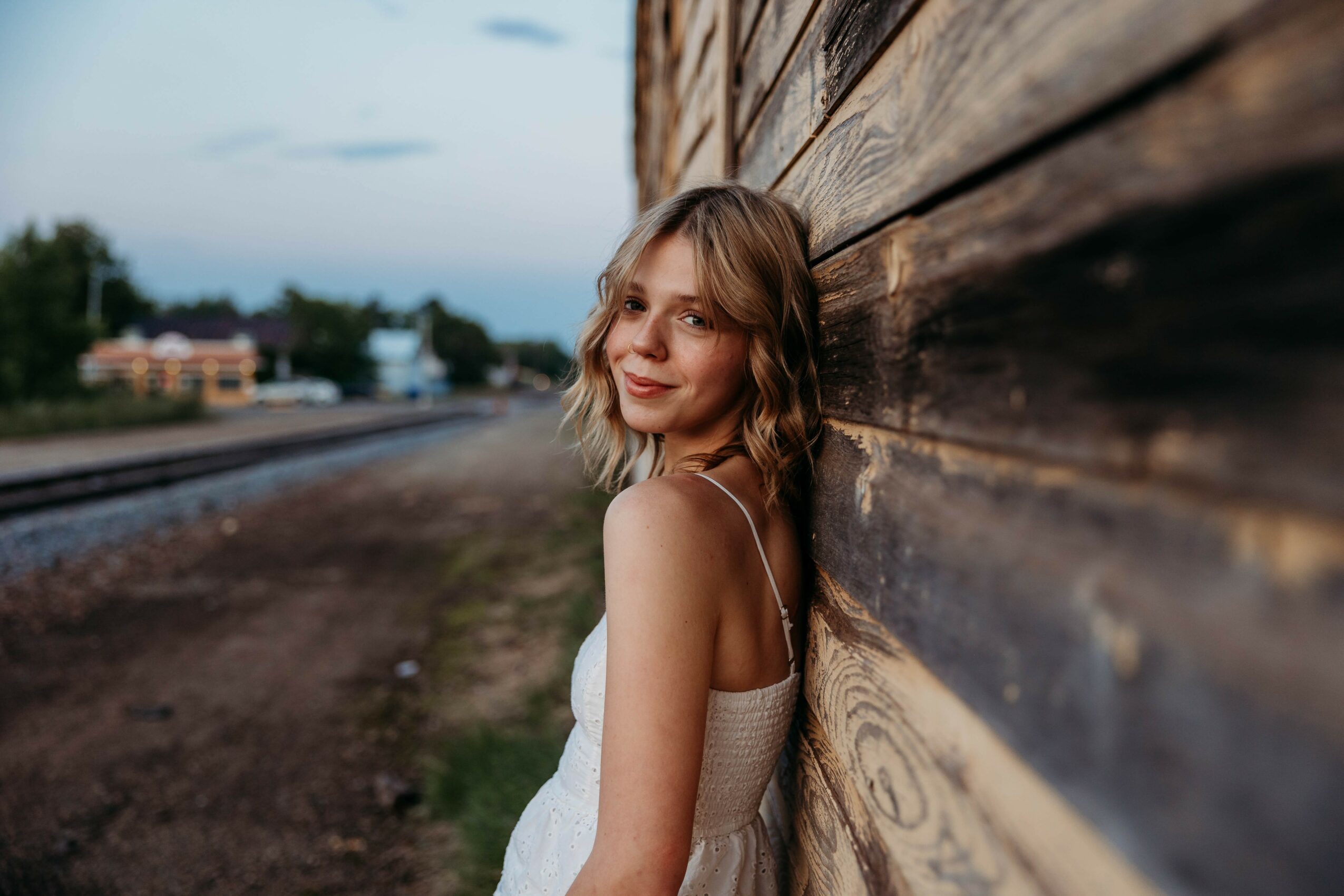 Young woman with shoulder-length hair leaning against a wooden wall outdoors, smiling, with a blurred background of trees and sky.
