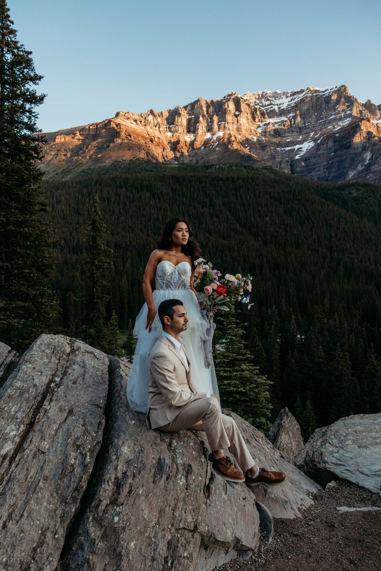 A bride and groom sit on rocks outdoors with mountains and a forest in the background, the bride holding a bouquet.