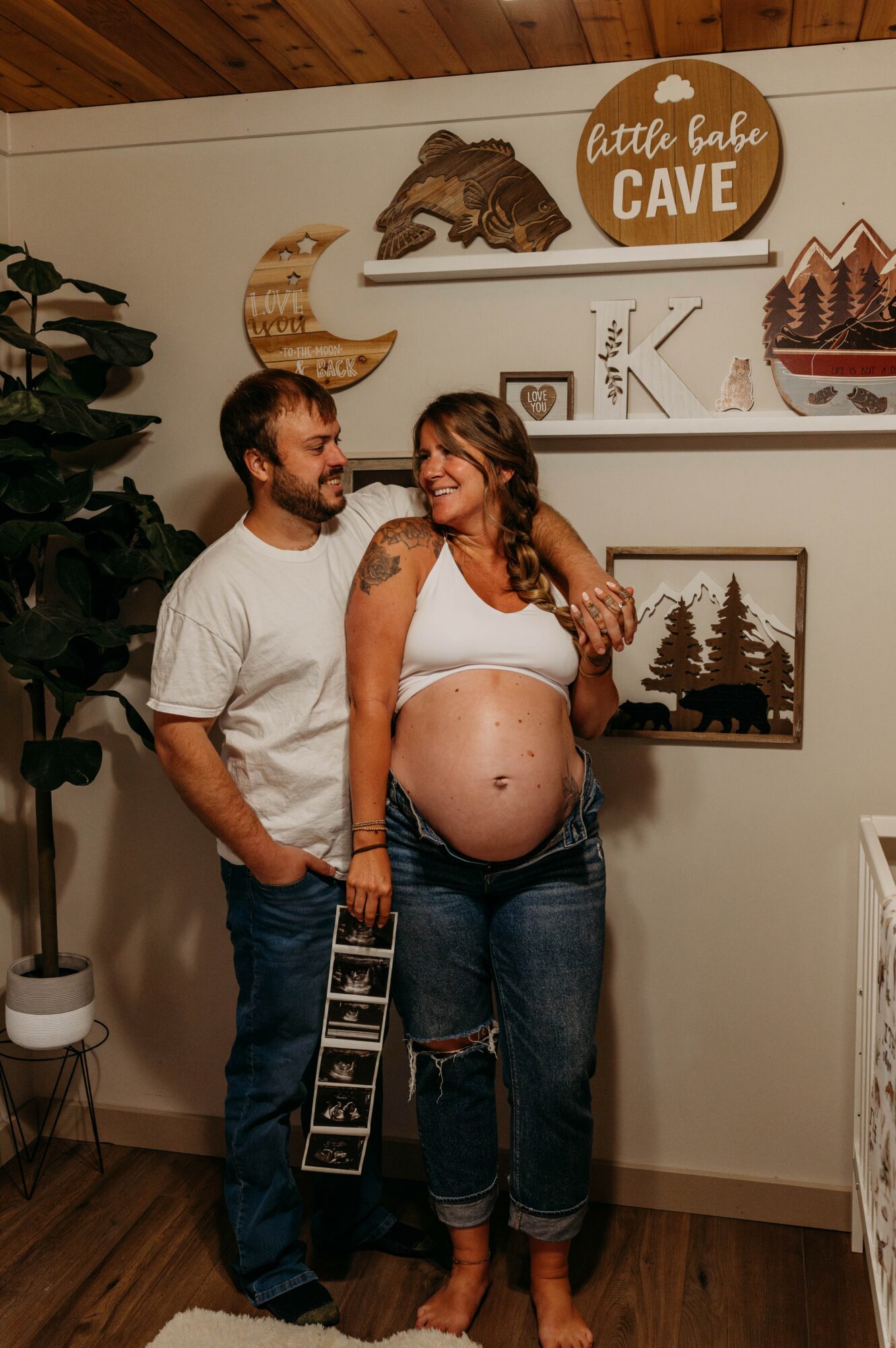 Pregnant woman with a tattoo on her arm and a man stand together in a room with wall decorations, smiling.