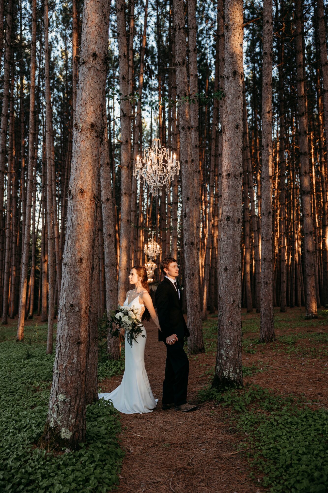 Bride and groom stand in a forest at sunset, holding hands, with a chandelier hanging among tall trees.