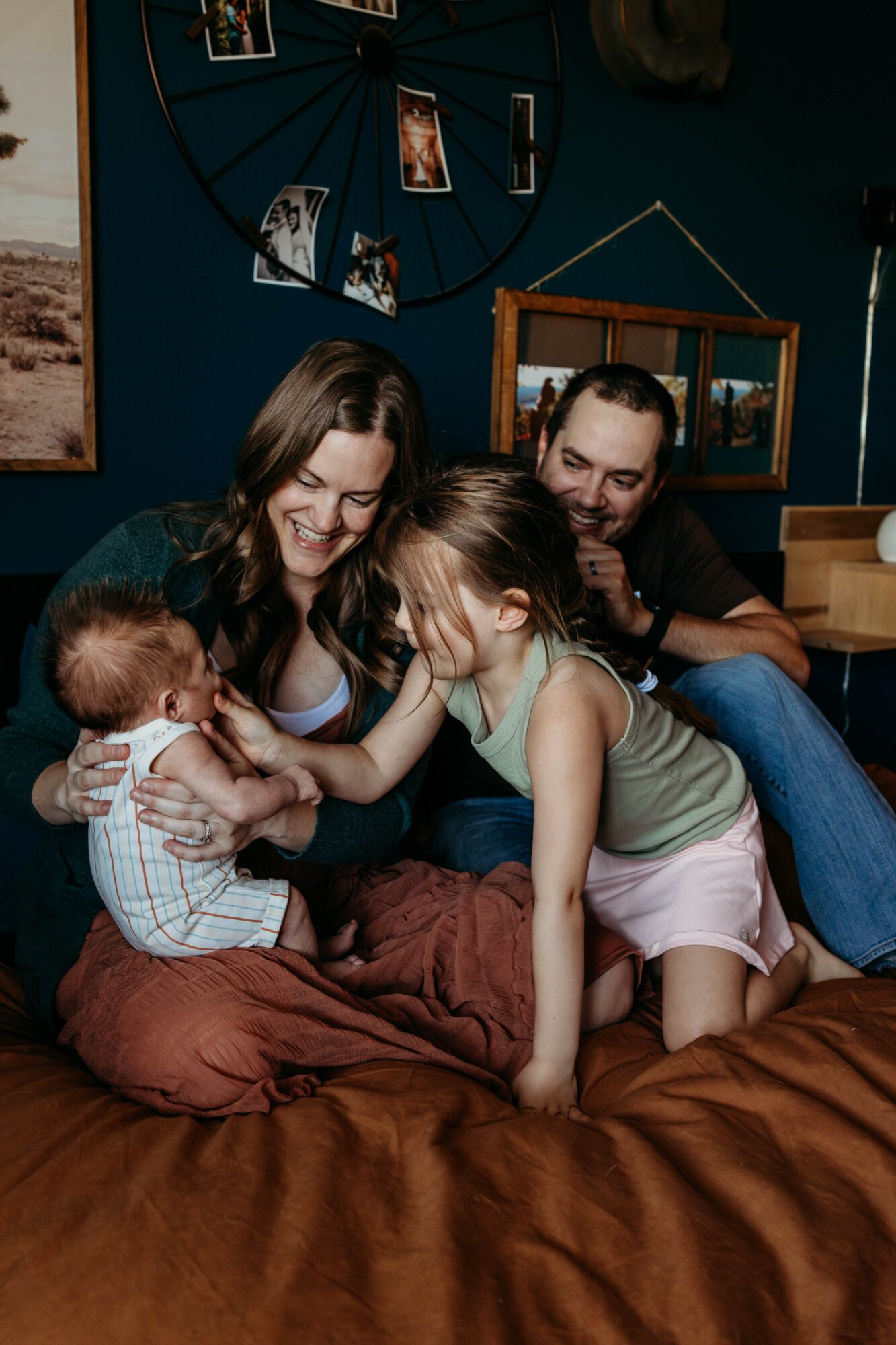 Family of four sitting on a bed, playing with a baby, smiling, in a cozy room with photos on the wall.