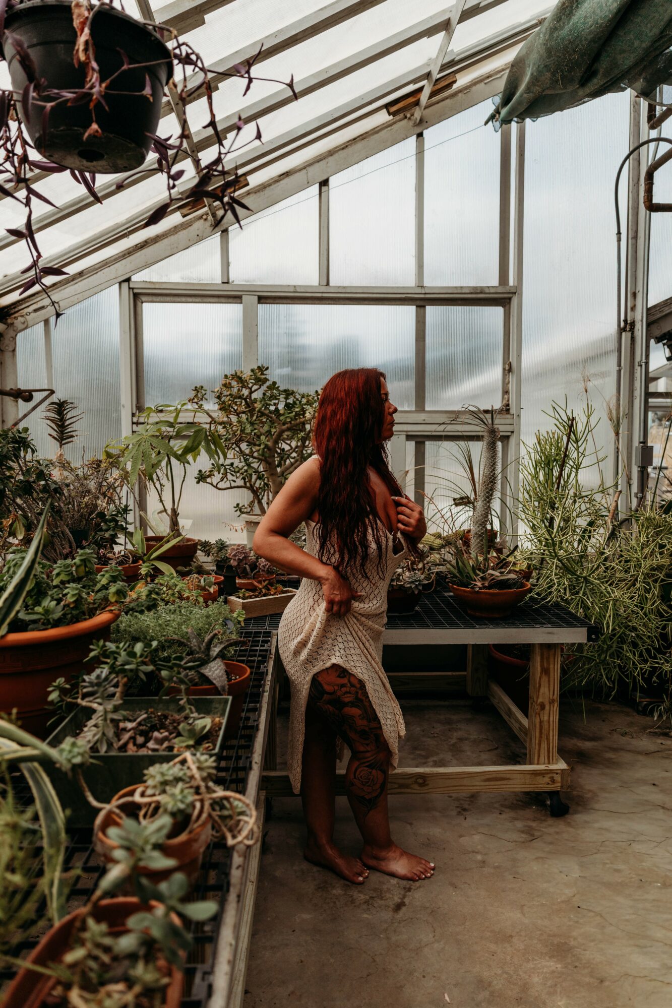Woman with long hair in a greenhouse surrounded by plants, holding a basket, standing barefoot on concrete floor.