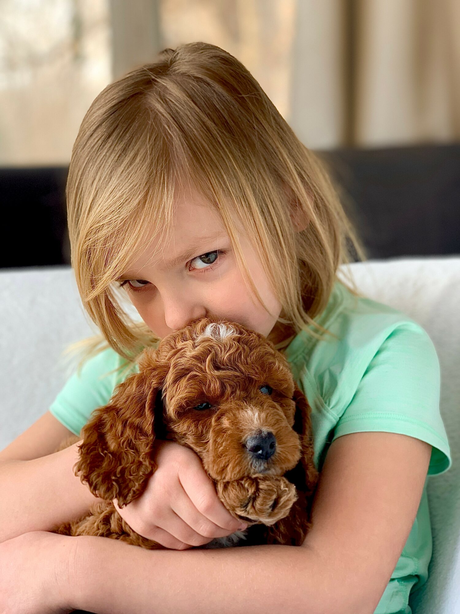 Young girl with blonde hair hugging a brown puppy indoors, smiling gently.