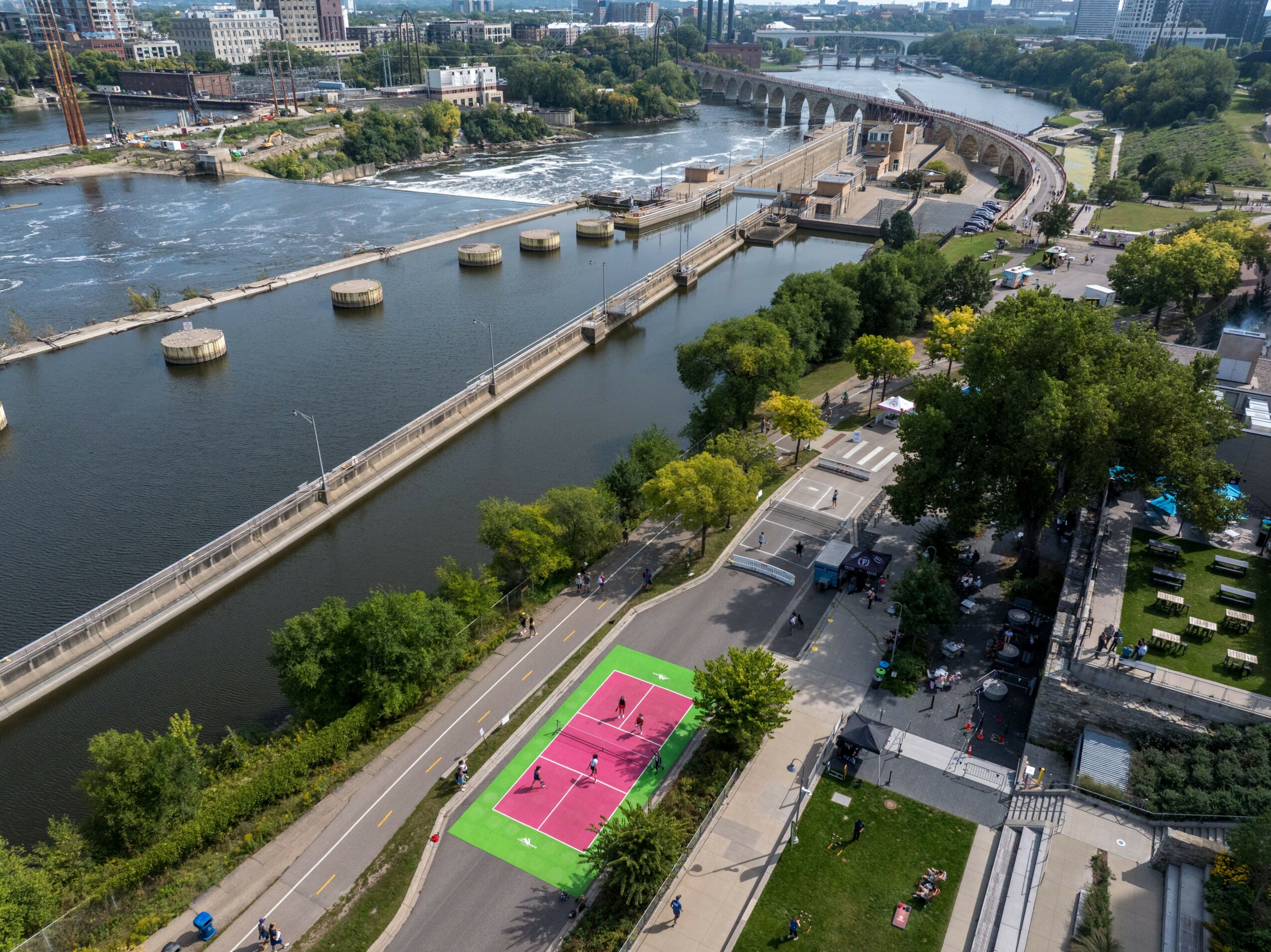 A river with a bridge and a pink and green sports court surrounded by trees and parking lots.
