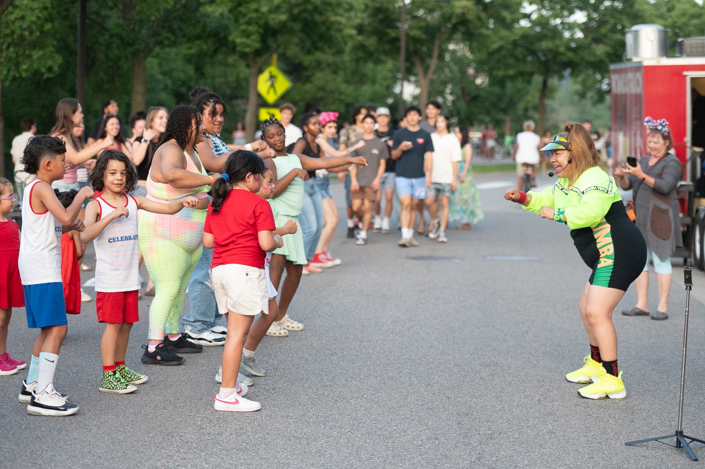 Group of children and adults outdoors, with a woman in bright clothing talking to children on a street.