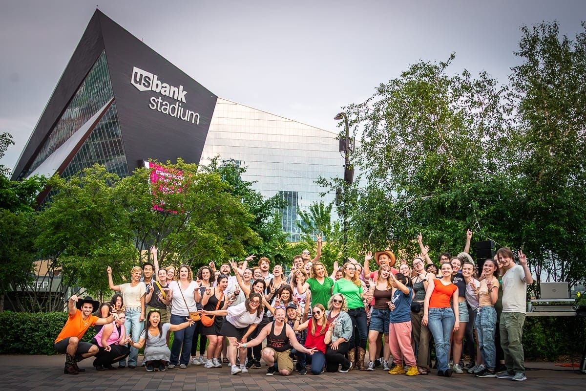 Group of people gathered outdoors in front of US Bank Stadium, with trees and the stadium visible in the background.