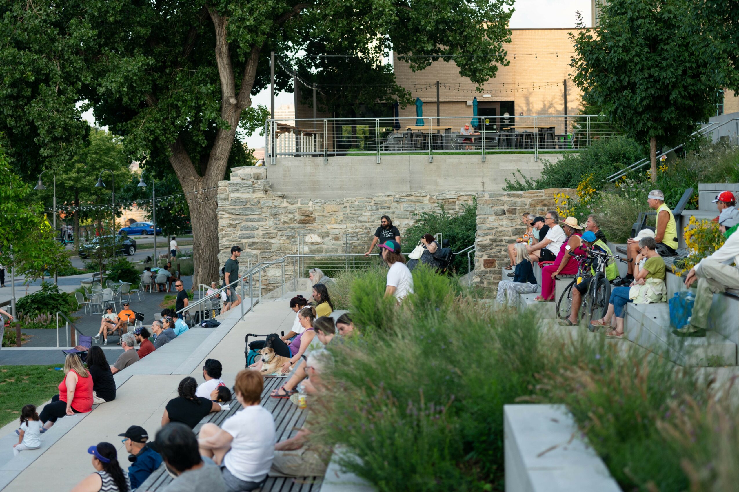 People sitting on outdoor steps and benches in a park with trees and greenery, some walking or standing, in daytime.