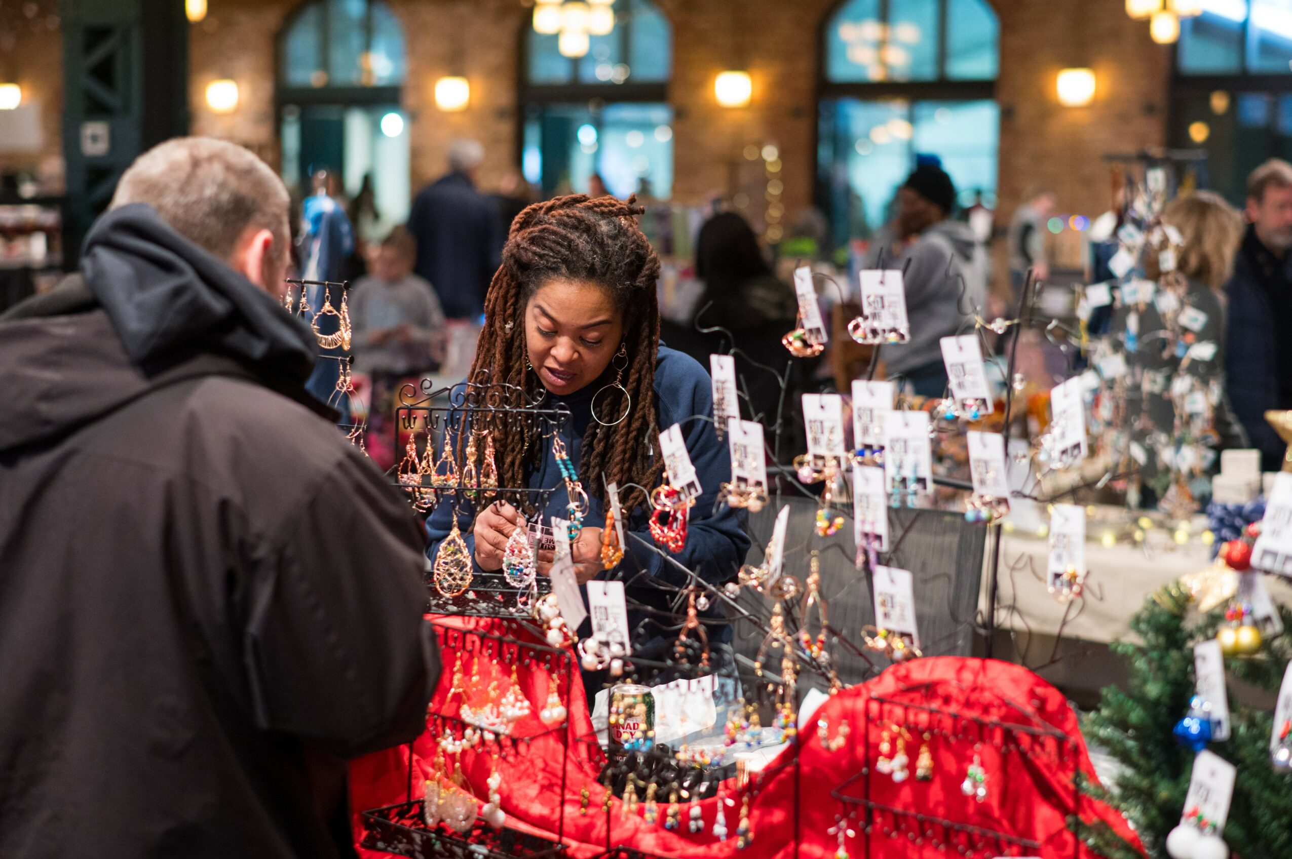 Woman with braided hair shopping at an indoor market stall with jewelry and accessories, surrounded by people and displays.