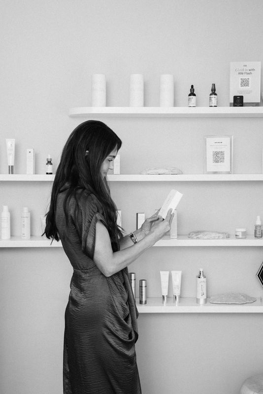 Woman with long dark hair reading a book in a minimalist room with shelves holding bottles and boxes