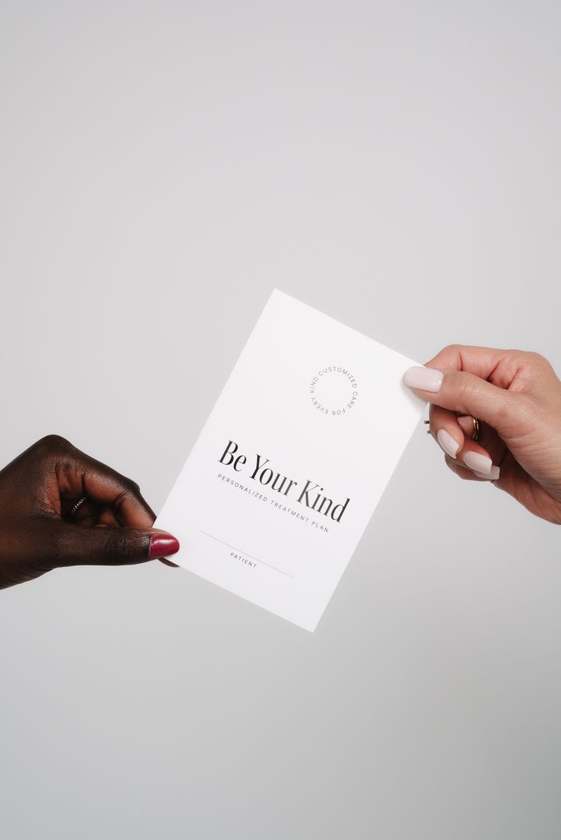 Two hands exchange a white card with the message 'Be Your Kind' against a plain background.