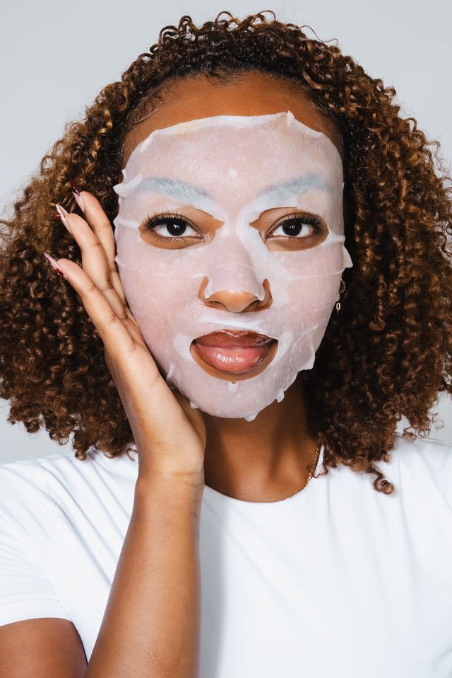 Woman with curly hair applying a facial mask, touching her face gently, wearing a white shirt, against a plain background.