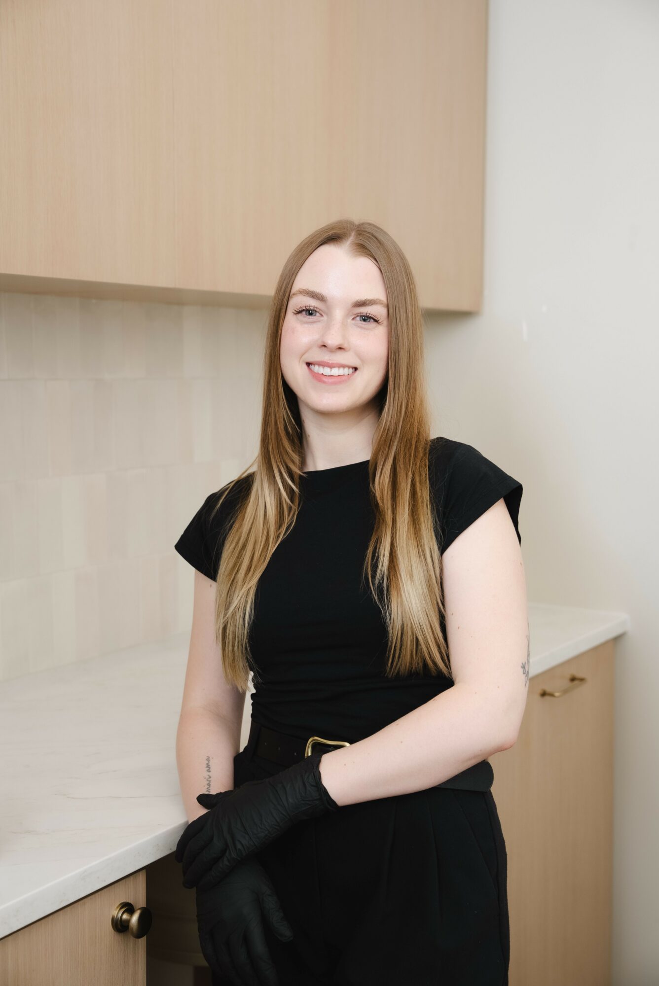Young woman with long hair smiling, wearing a black dress and gloves, standing indoors near a cabinet.