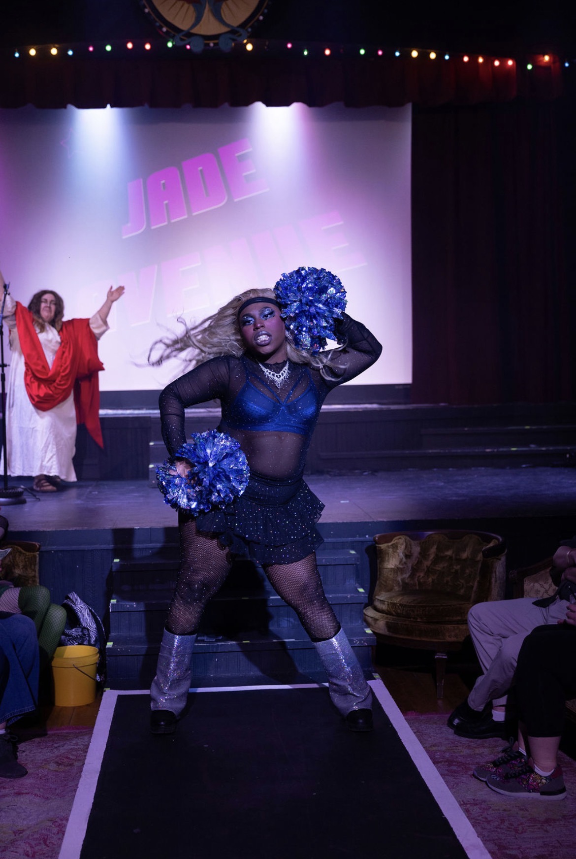 Performer on stage with blue pom-poms, wearing a black outfit, in front of a lit backdrop with purple letters, audience watching.