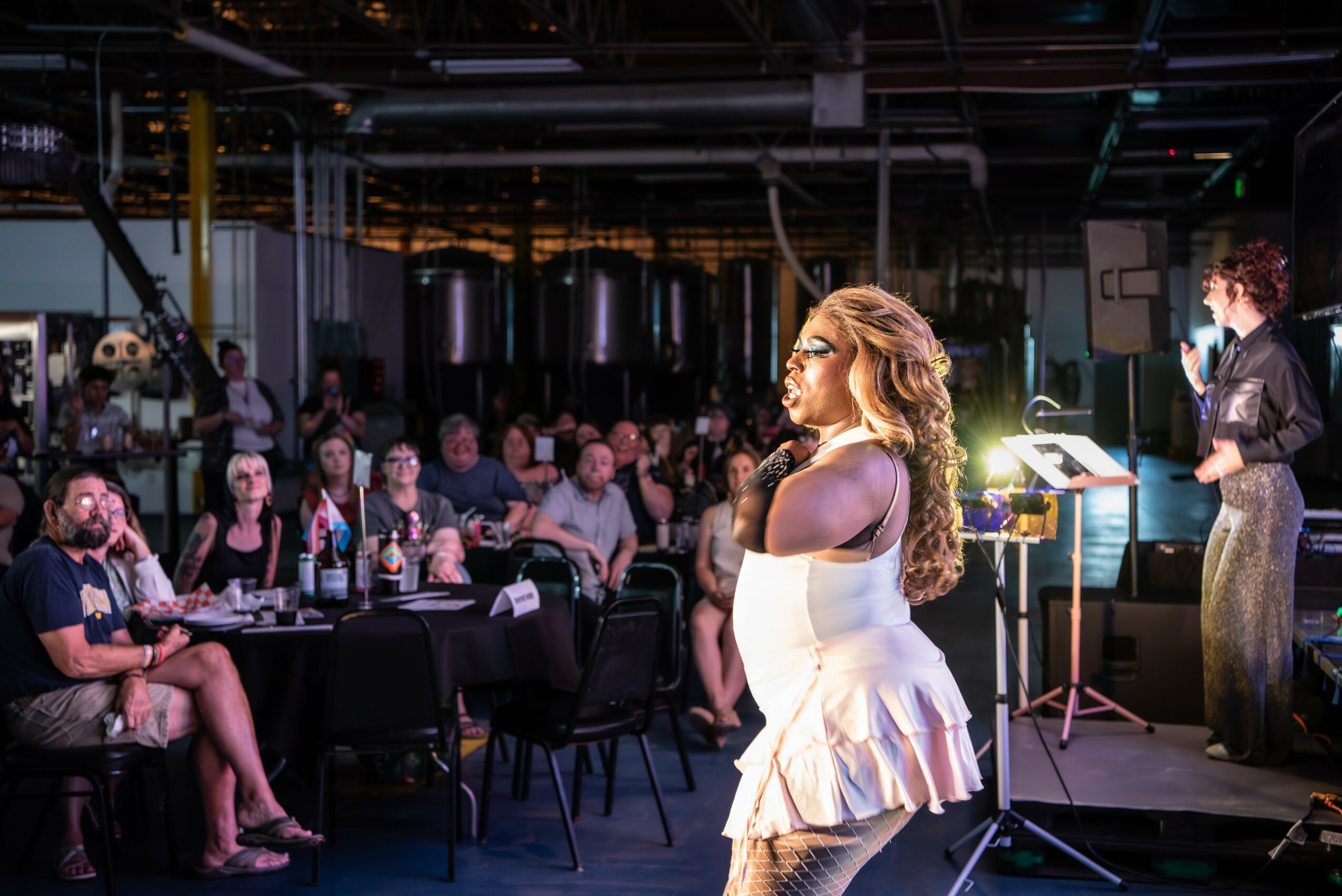 A woman in a white dress stands on stage speaking to an audience in a large event space.