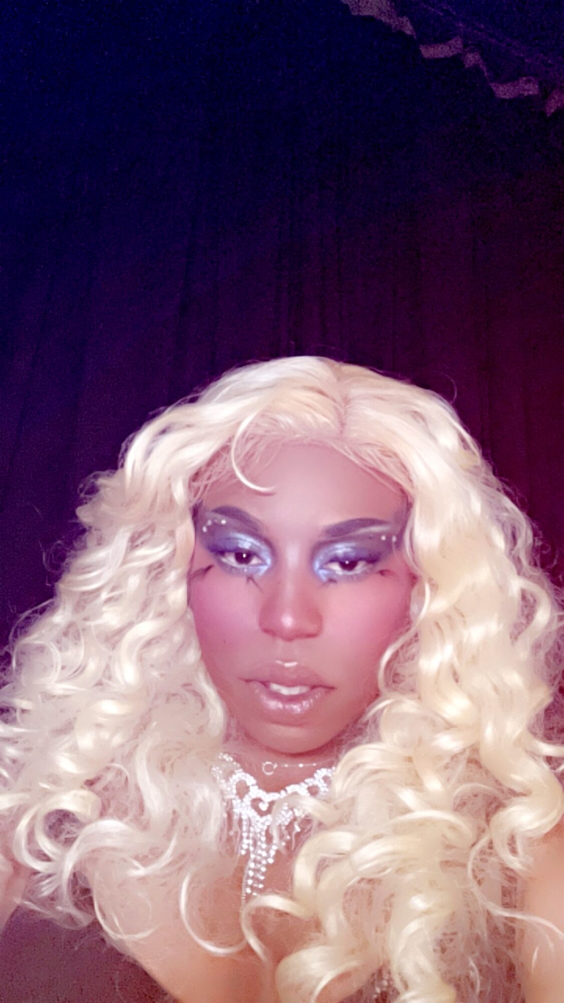 Person with long, curly platinum blonde hair and dramatic makeup, wearing a necklace, against a dark background.