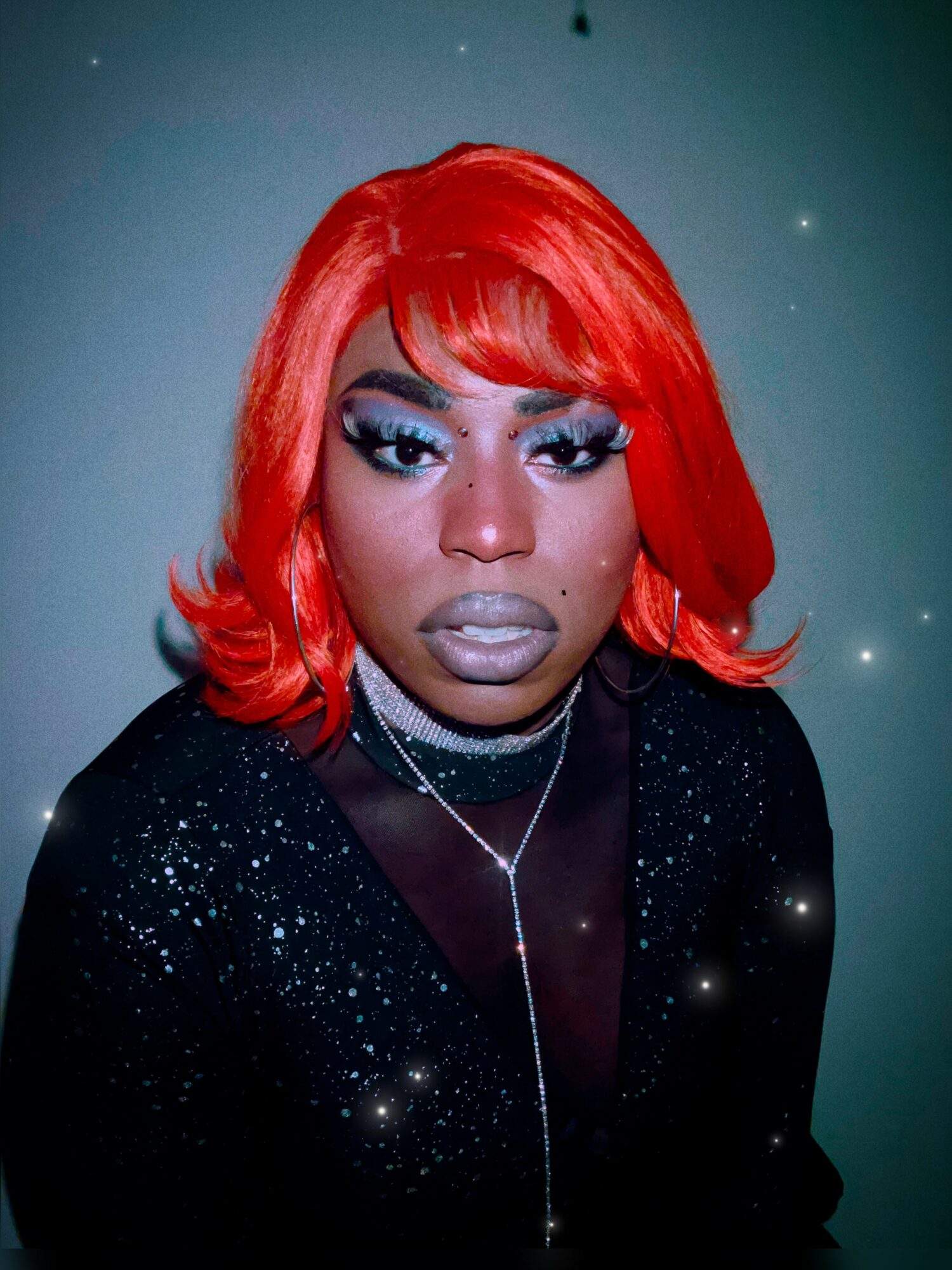 Person with bright red hair, bold makeup, wearing a black sparkly top and a necklace, looking at the camera.