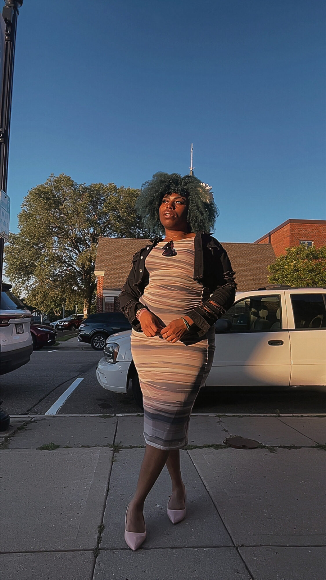 Woman standing on sidewalk with cars and trees in background, clear blue sky, sunlight, wearing a striped dress and pink shoes.