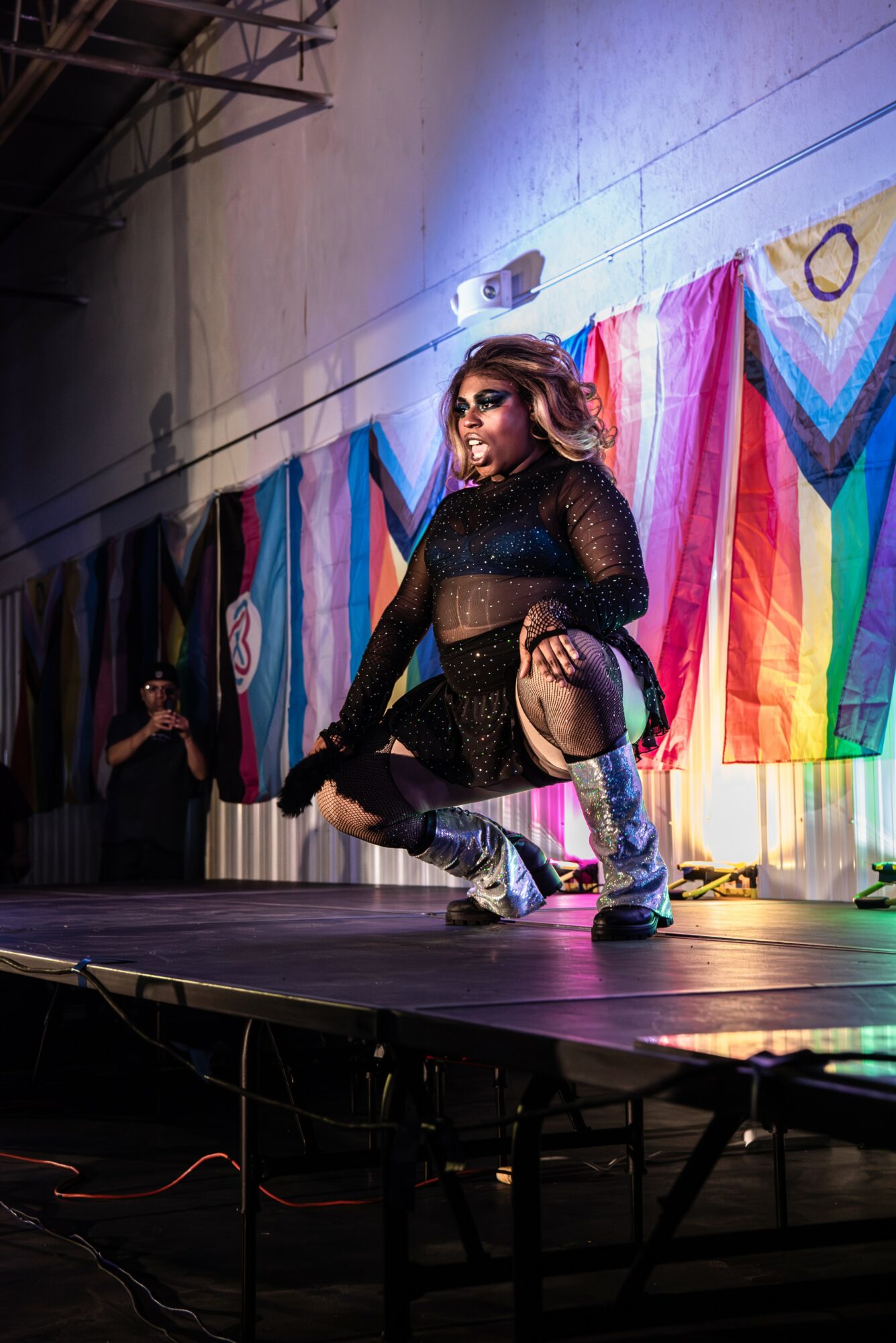 Performer crouching on stage with colorful rainbow flags in background, wearing black outfit and sunglasses.