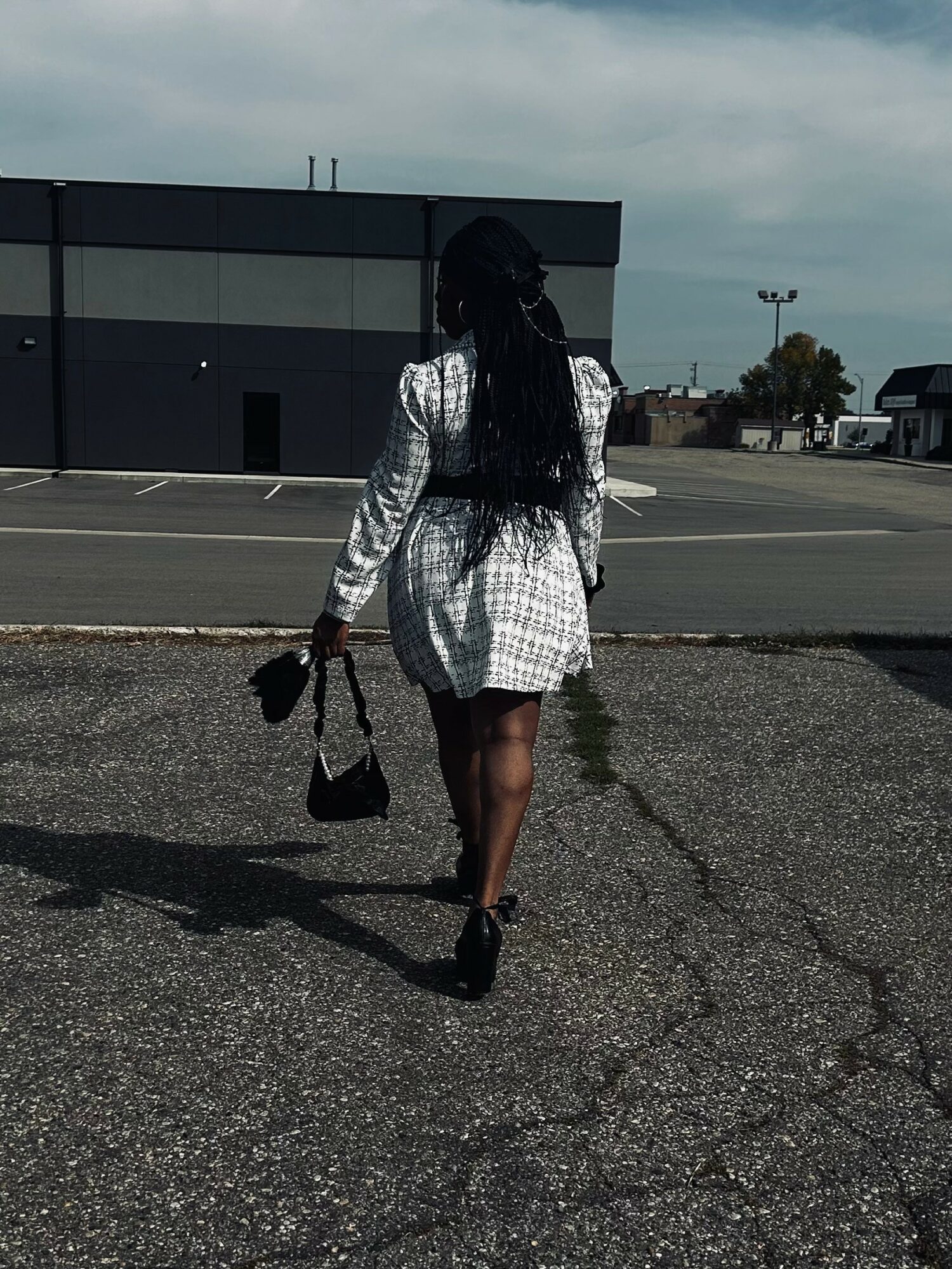 Person walking away in a parking lot, wearing a white dress, carrying a bag, with long dark hair, under a cloudy sky.