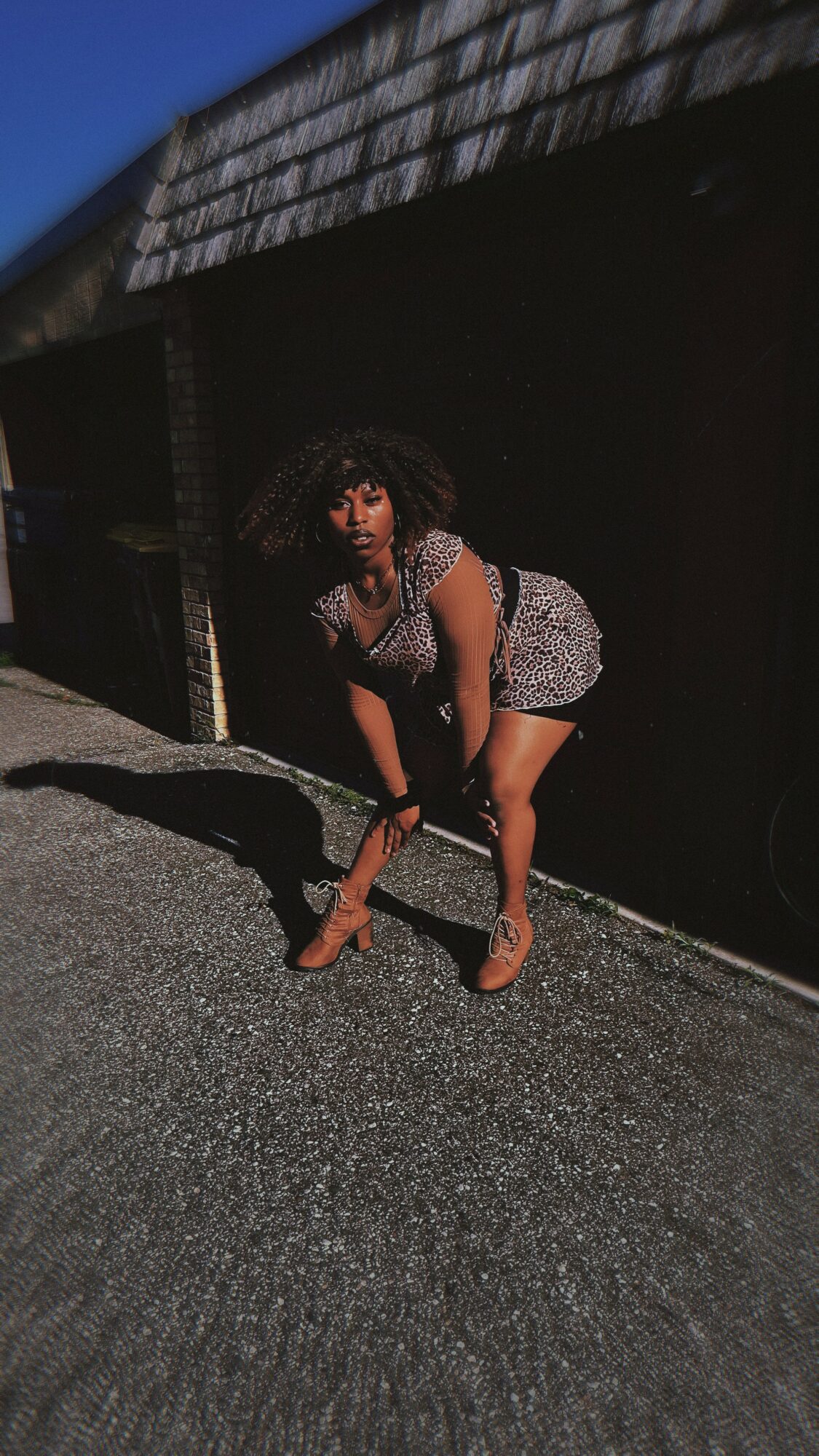 Person crouching on asphalt ground in front of a dark building, wearing a patterned dress and sandals.