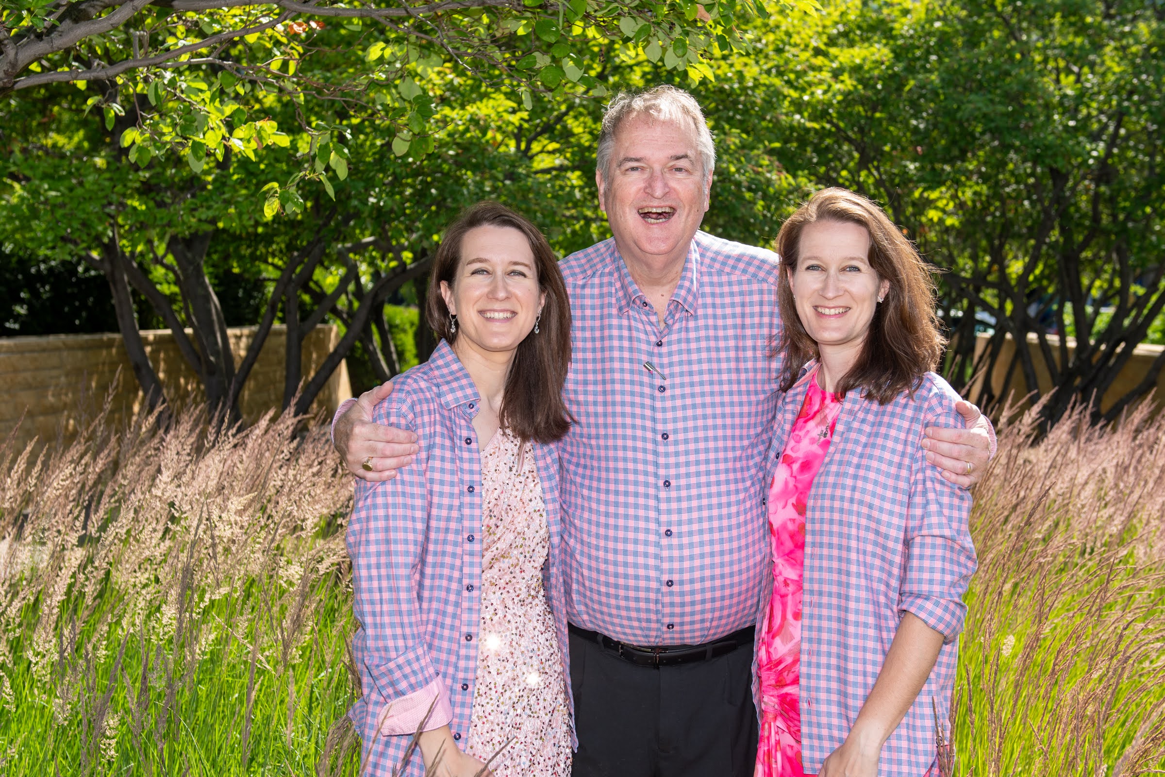 Three people smiling outdoors in a grassy area with trees, standing close together with arms around each other.