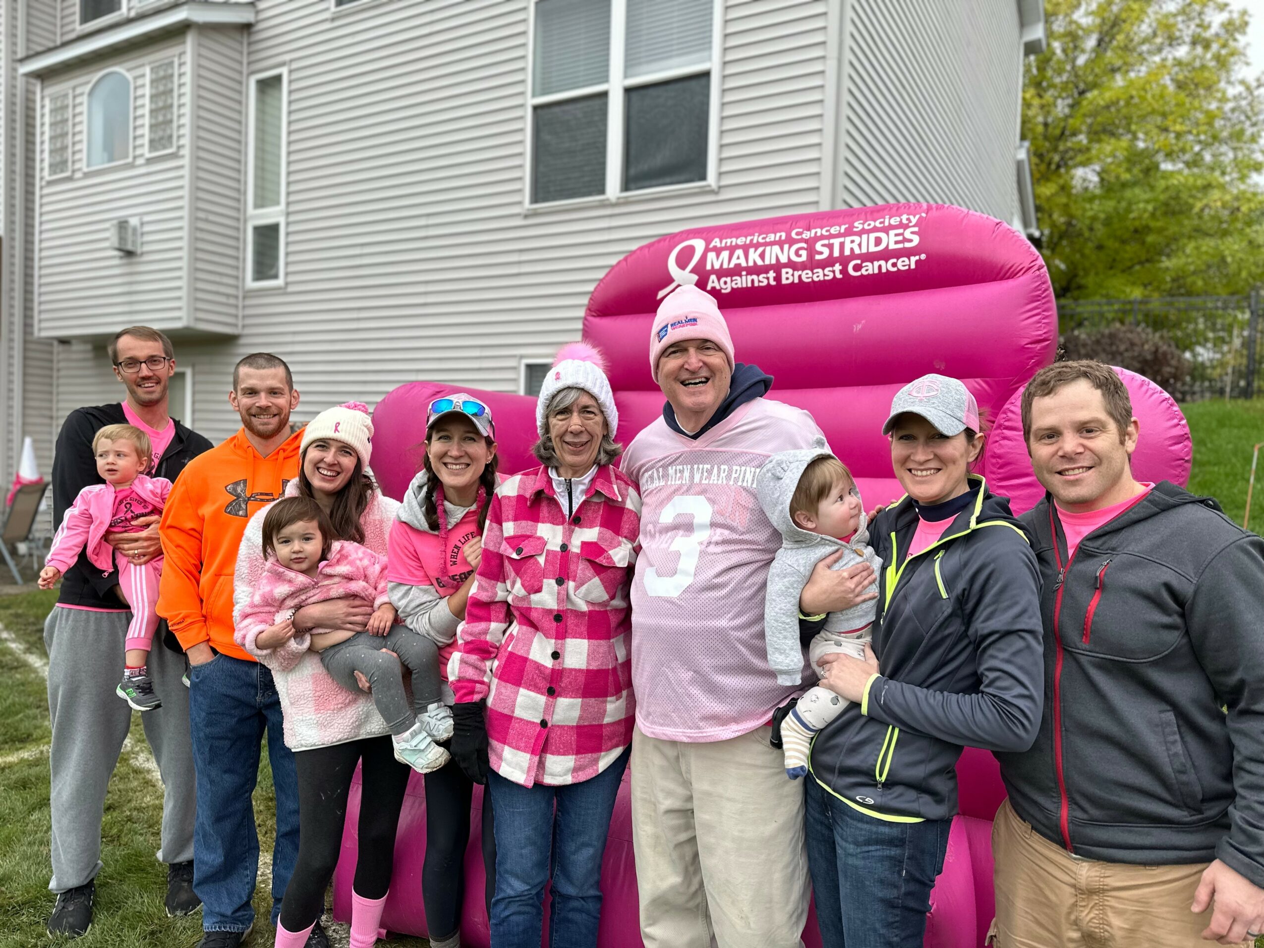 Group of people standing outdoors in front of a house, some holding children, with a pink breast cancer awareness float behind them.