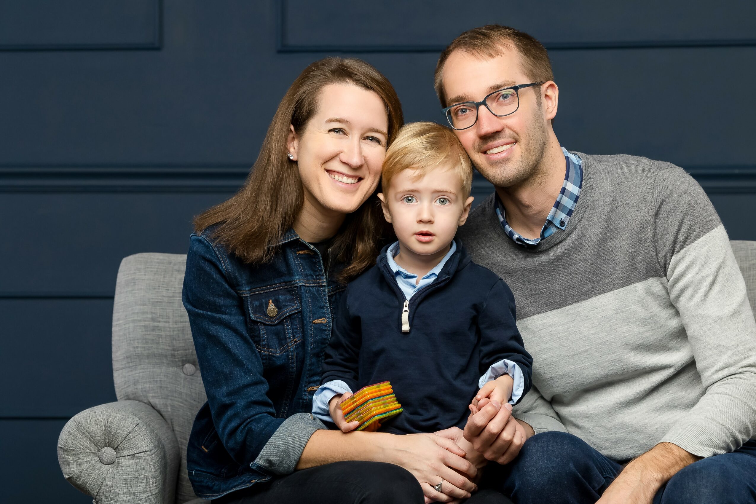 Family of three sitting on a gray couch, smiling, with a dark blue wall background.