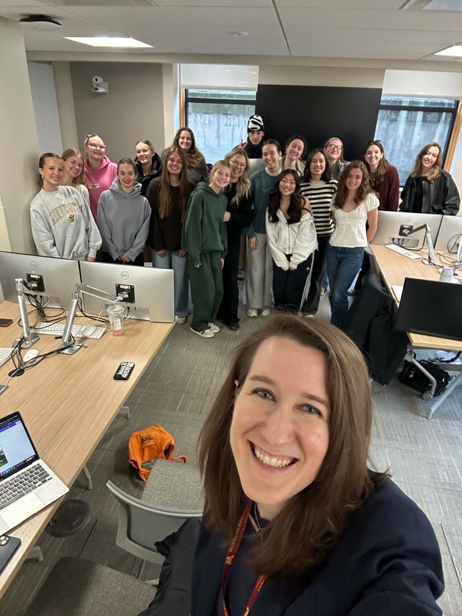 Group of people in a conference room, smiling and posing for a photo, with a woman taking selfie in foreground.