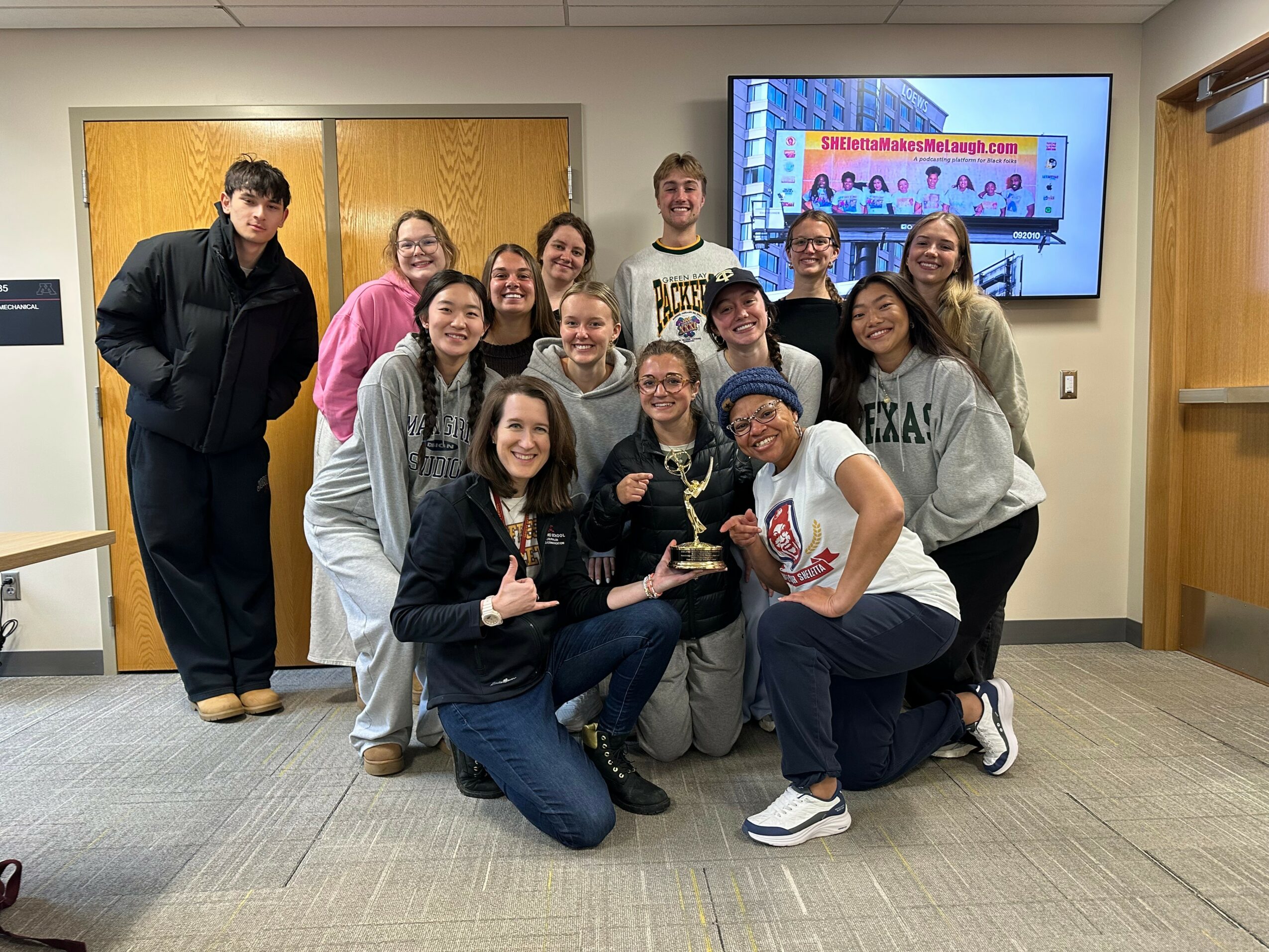 Group of 13 people, some holding trophies, posing in a room with a large screen displaying a website, smiling at the camera.