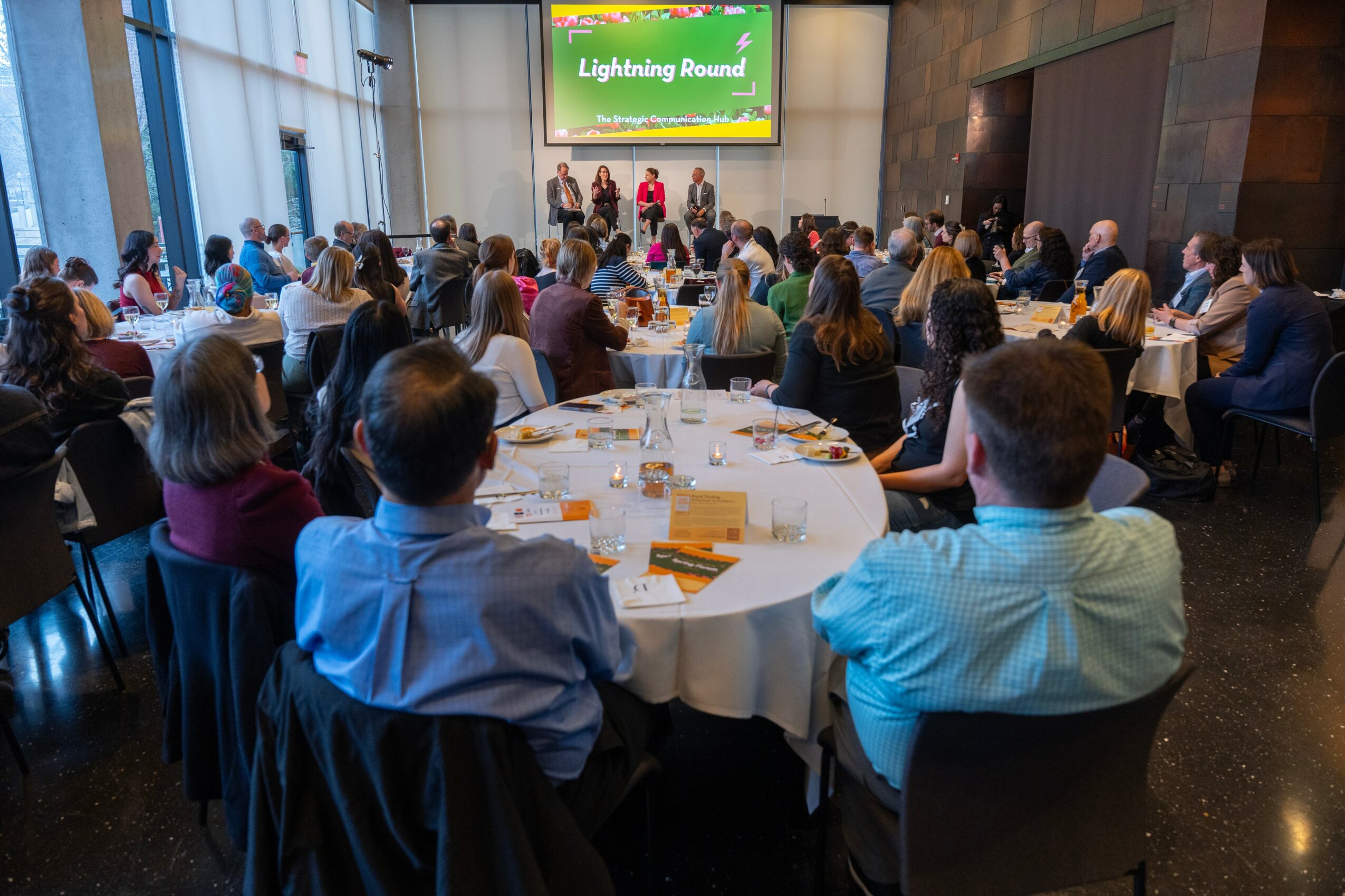 People seated at round tables watching a presentation on a large screen in a conference room.