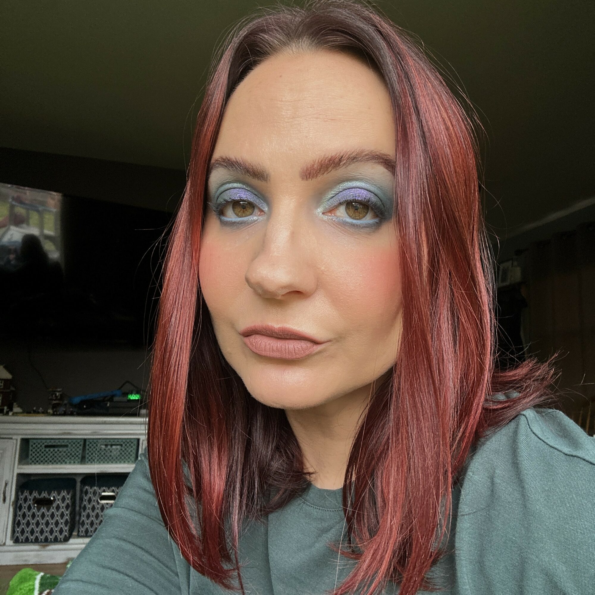 Woman with shoulder-length reddish-brown hair and blue eyeshadow, looking at the camera indoors.