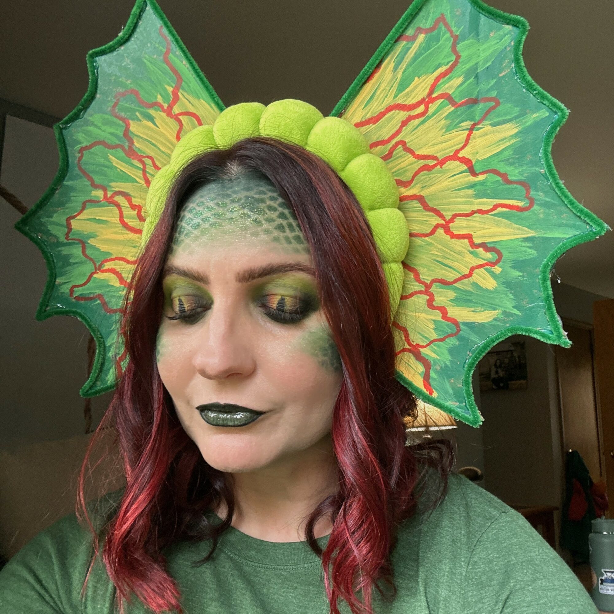 Person with green and yellow headpiece resembling a flower, green makeup, and dark lipstick, looking down.