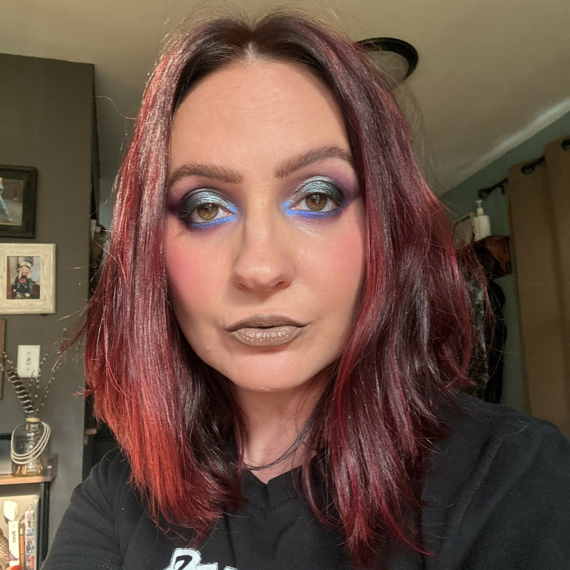 Woman with shoulder-length wavy hair, blue eyeshadow, and metallic lipstick, wearing a black top indoors.