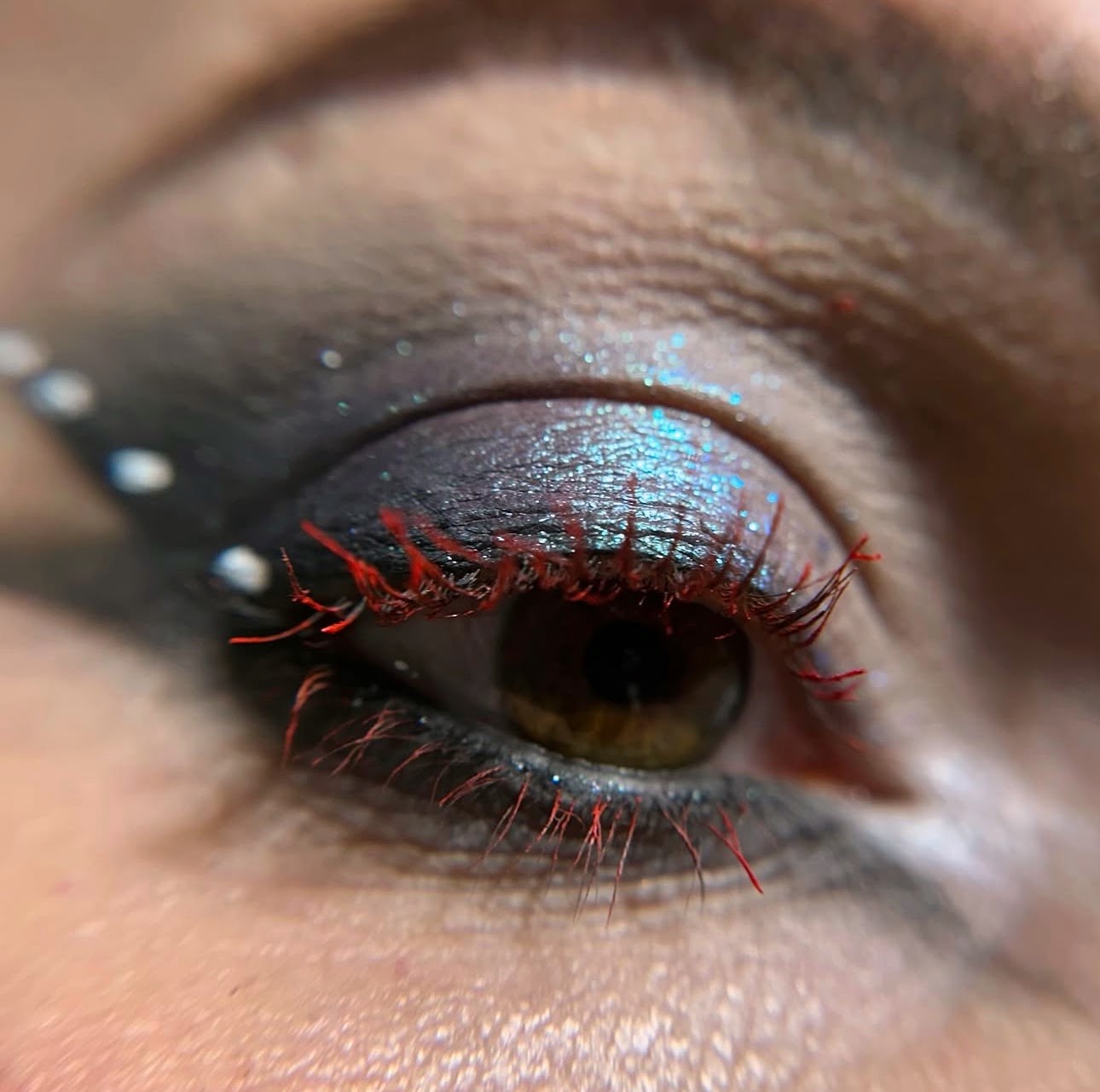 Close-up of a person's eye with colorful eyeshadow, red eyelashes, and small white dots on the eyelid.