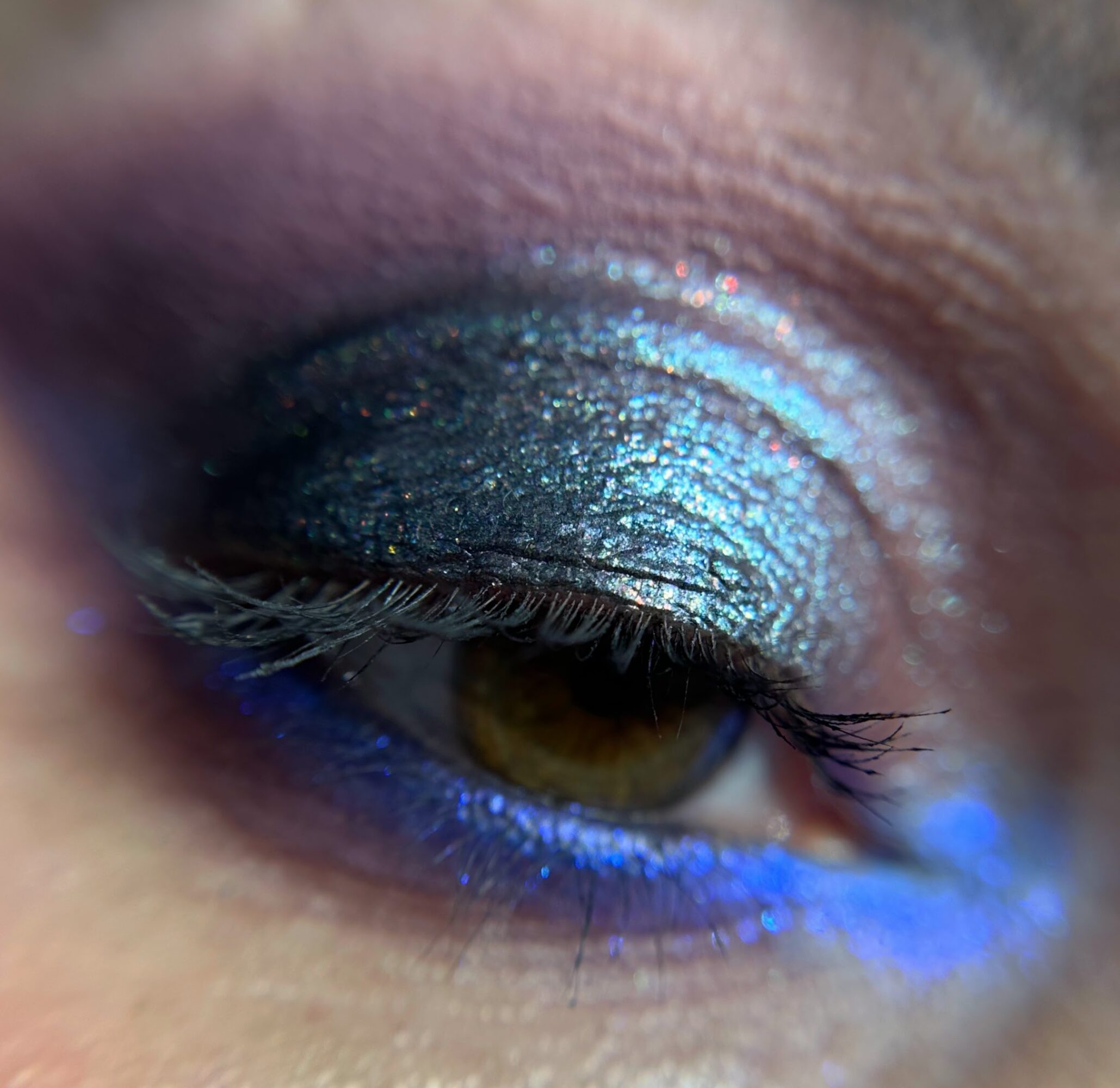 Close-up of a person's eye with shimmering blue and purple eyeshadow and eyeliner, and long eyelashes.