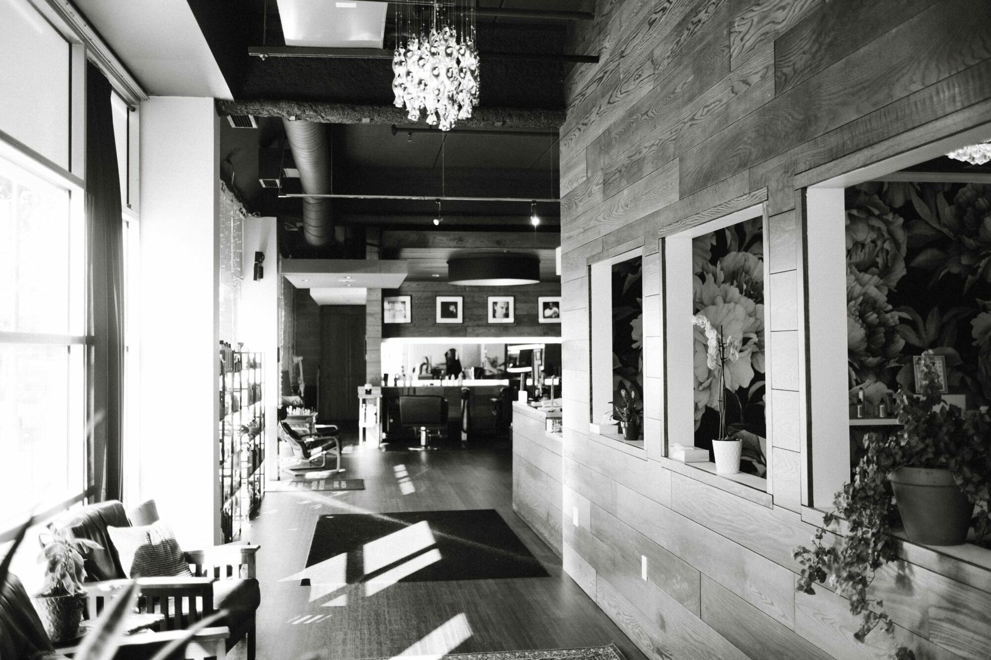 Interior of a modern cafe with large windows, wooden walls, and a chandelier, with sunlight casting shadows inside.