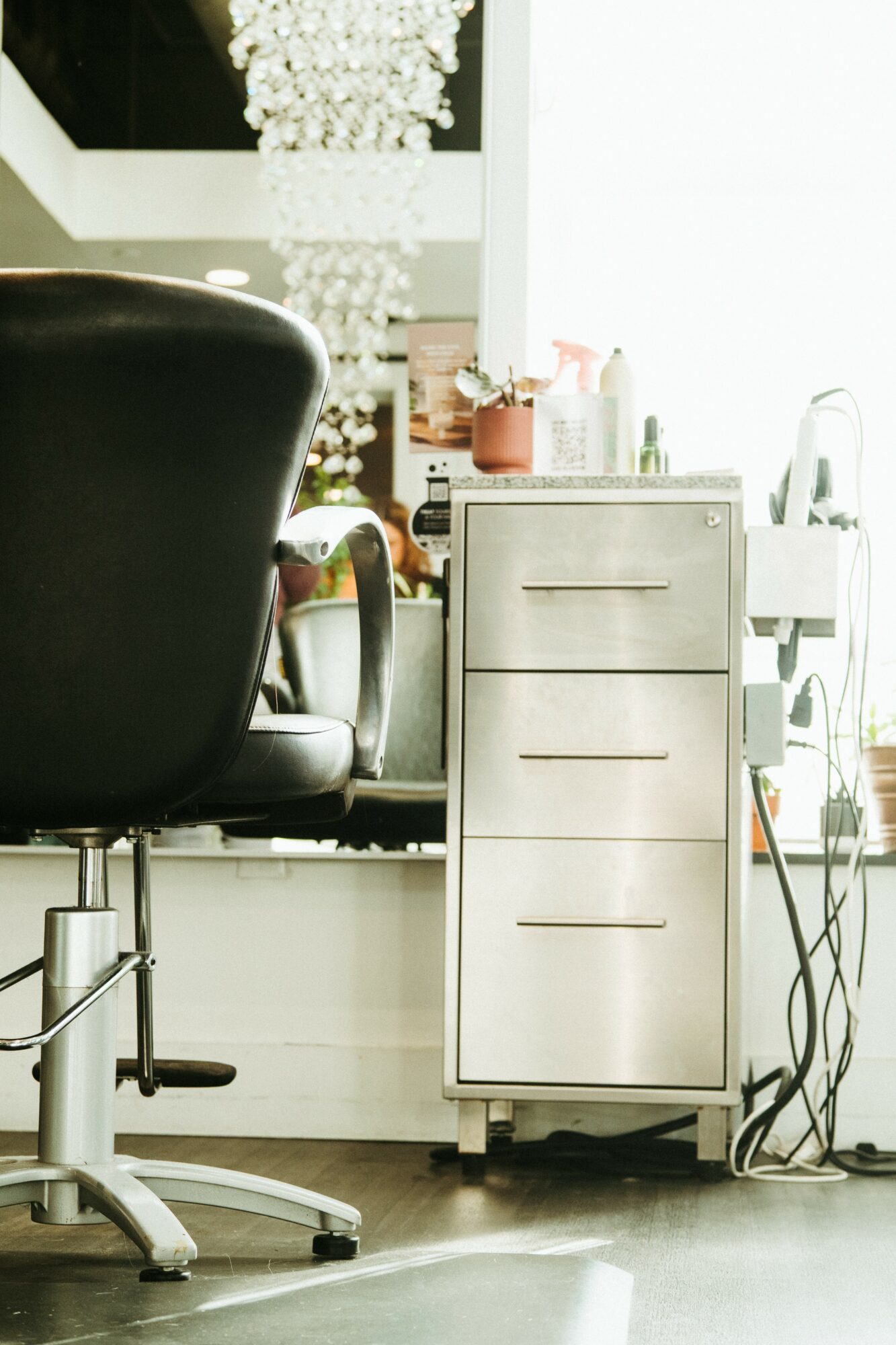 Office space with a black chair, white cabinet, and various items on top, near a window with bright light.