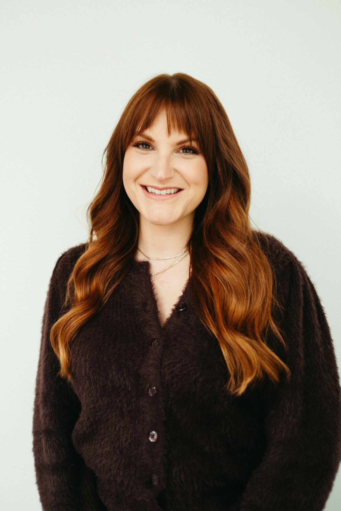 Smiling woman with long, wavy, reddish-brown hair wearing a dark sweater against a plain light background.